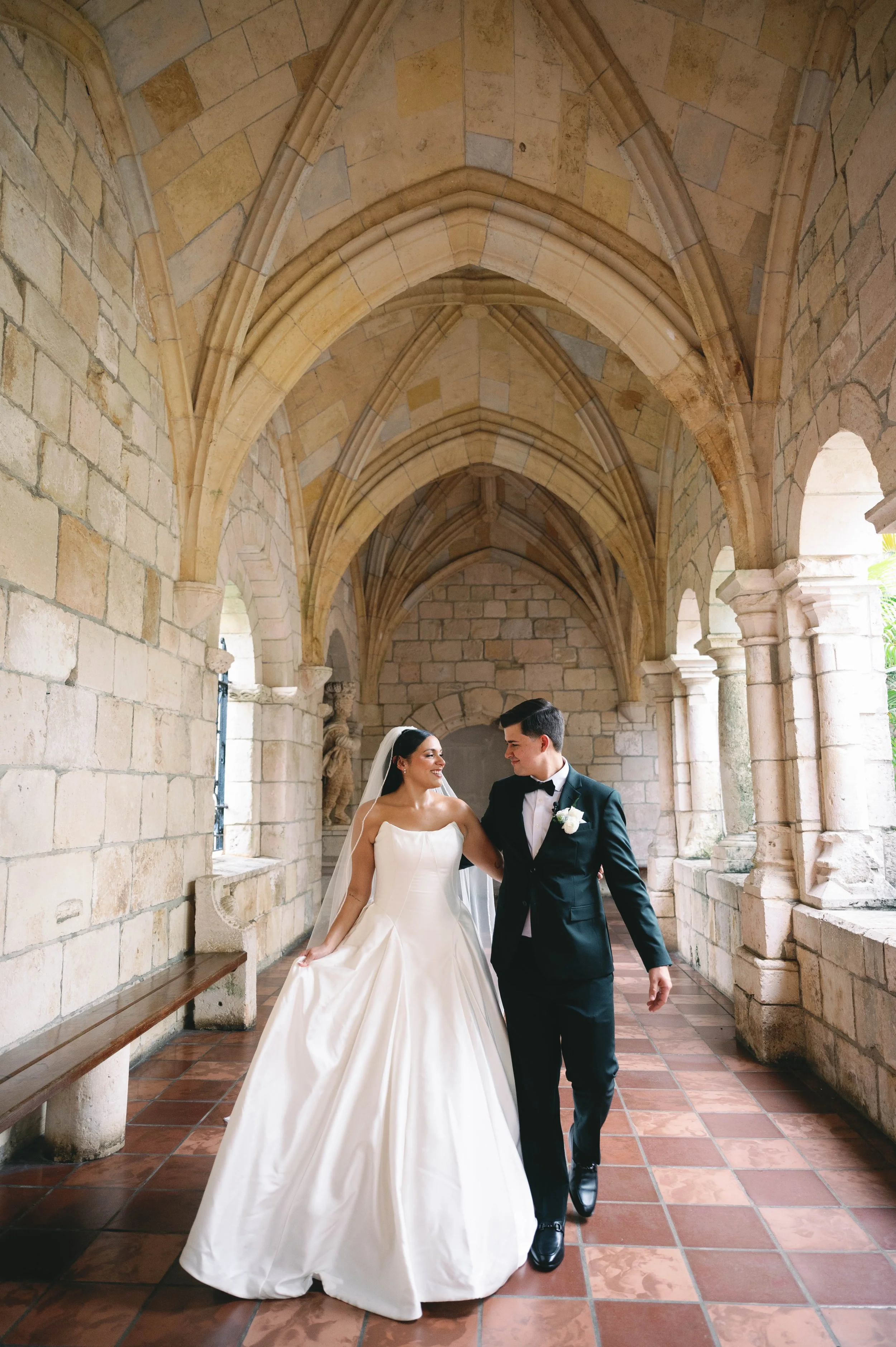 A bride and groom walking together in a stone archway with vaulted ceilings, the bride in a white wedding dress and the groom in a black tuxedo with a bowtie.