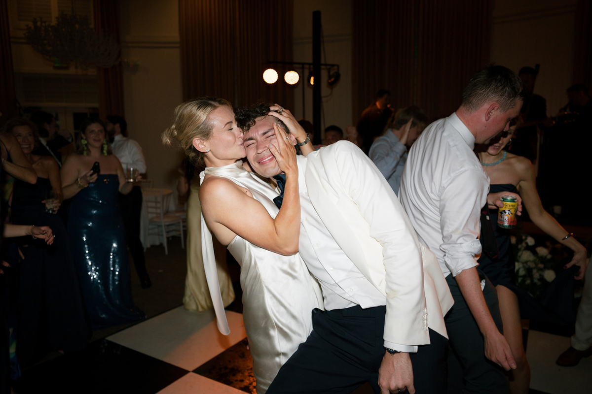 A woman in a satin dress and a man in a white suit share a kiss at a party, with the woman holding the man's face and the man smiling with eyes closed. People are dancing and socializing in the background.