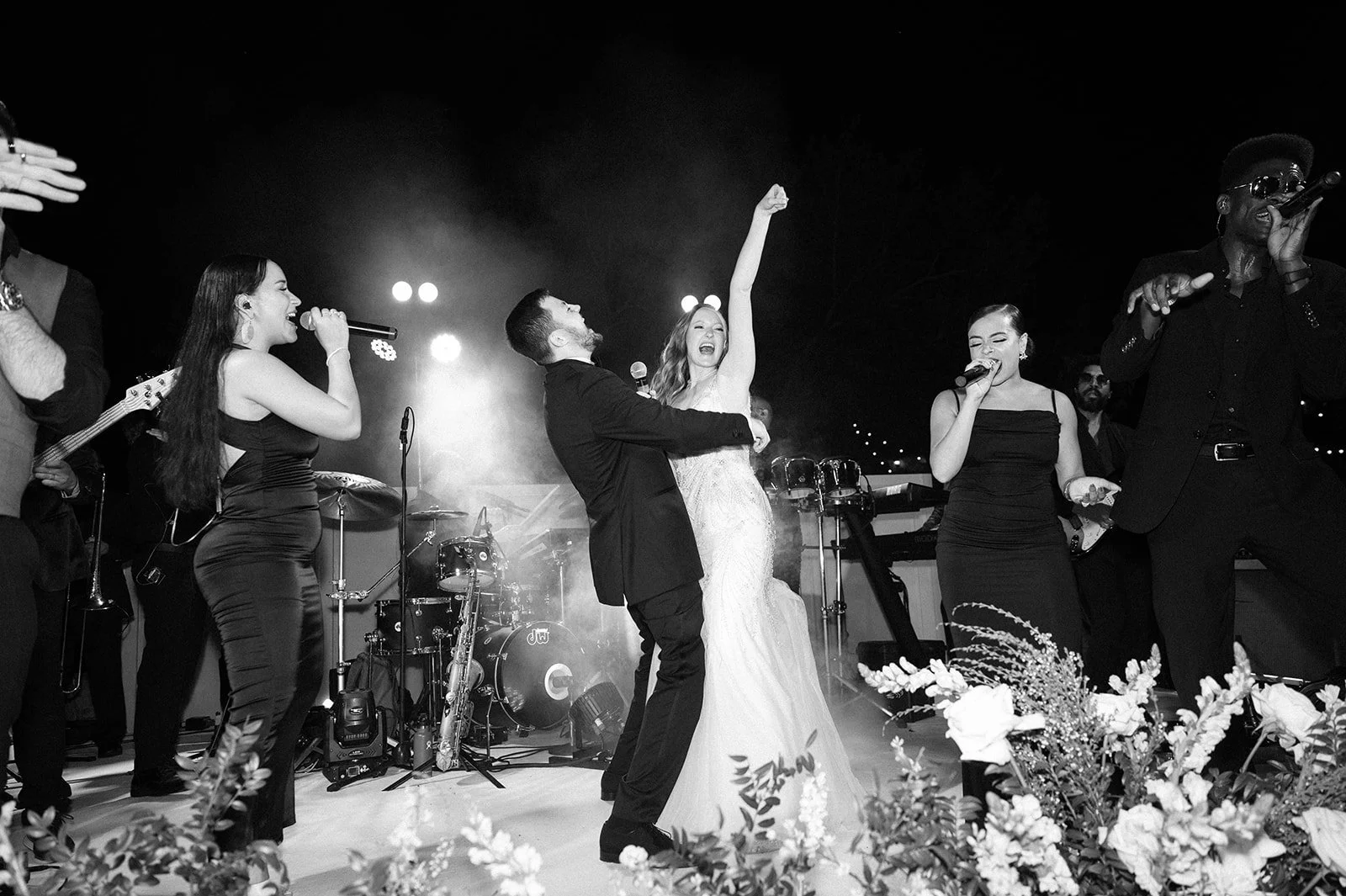 Black and white photo of a wedding celebration with a band performing. The bride and groom are dancing happily in the center, surrounded by musicians singing and playing instruments, with floral decorations in the foreground.