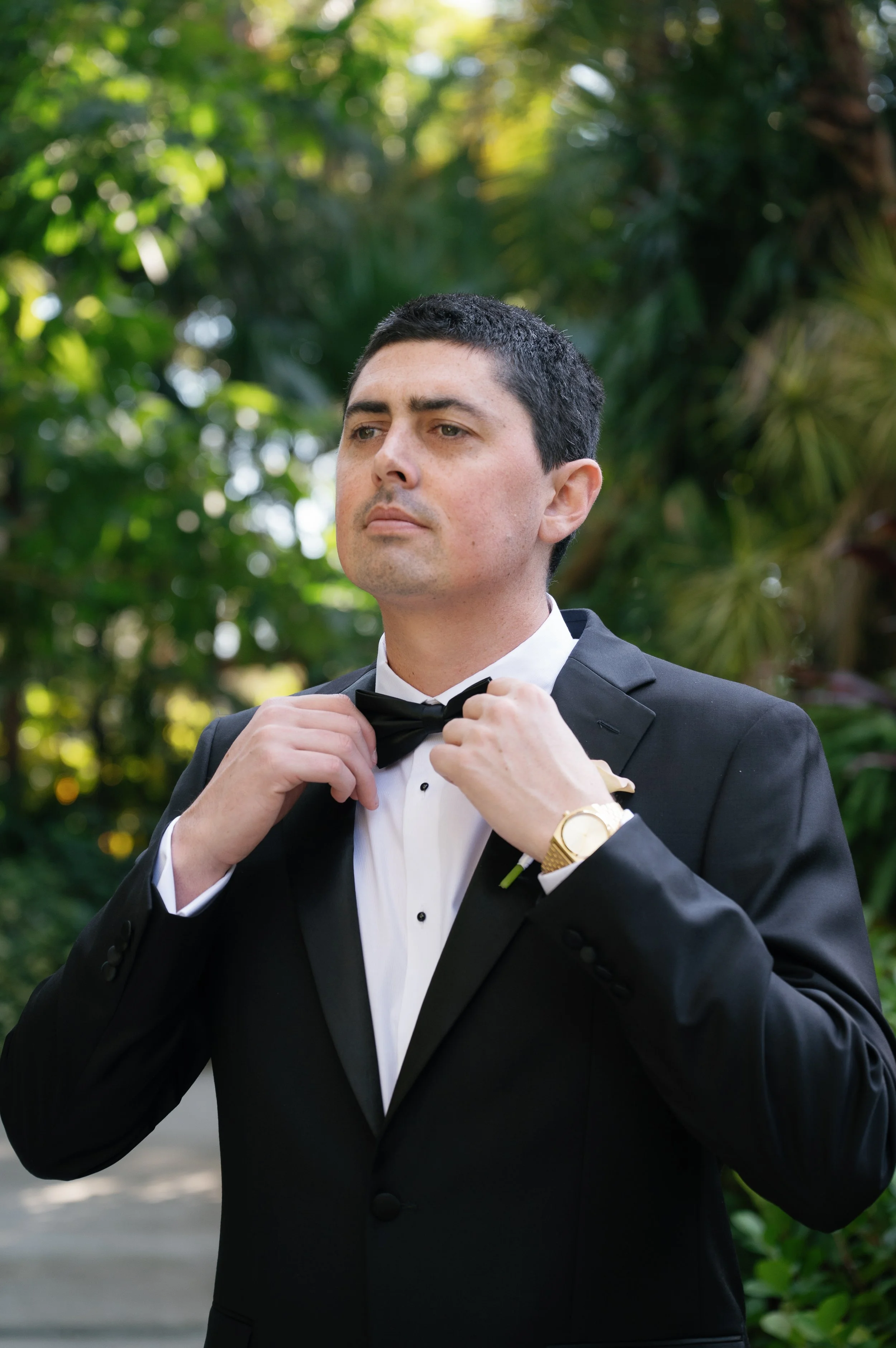 A man in a tuxedo adjusting his bowtie outdoors surrounded by green foliage.