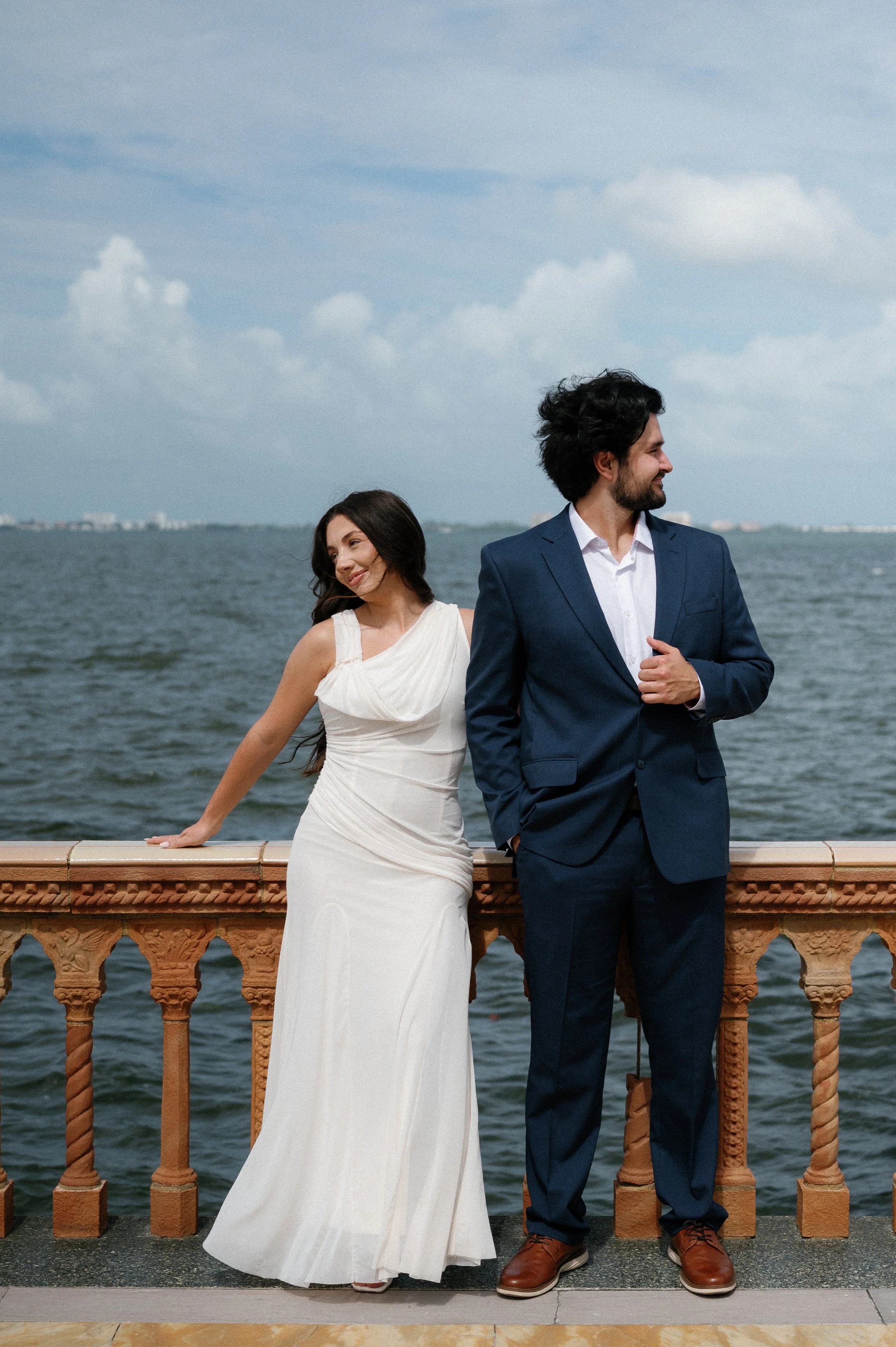 A woman in a white dress and a man in a dark blue suit stand by a railing near water, with a cloudy sky in the background.