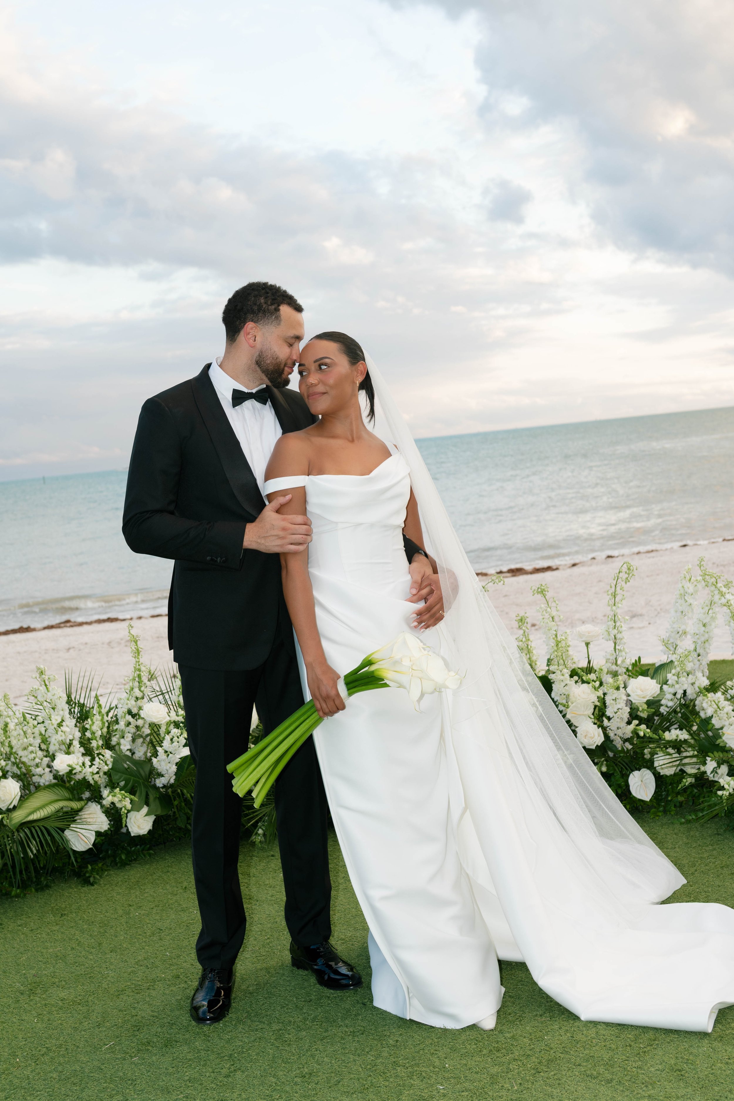 A bride and groom stand close together on a beach wedding, with the ocean in the background. The bride wears a white off-the-shoulder wedding gown and holds a bouquet of white calla lilies, while the groom wears a black tuxedo with a bow tie. They look at each other lovingly.