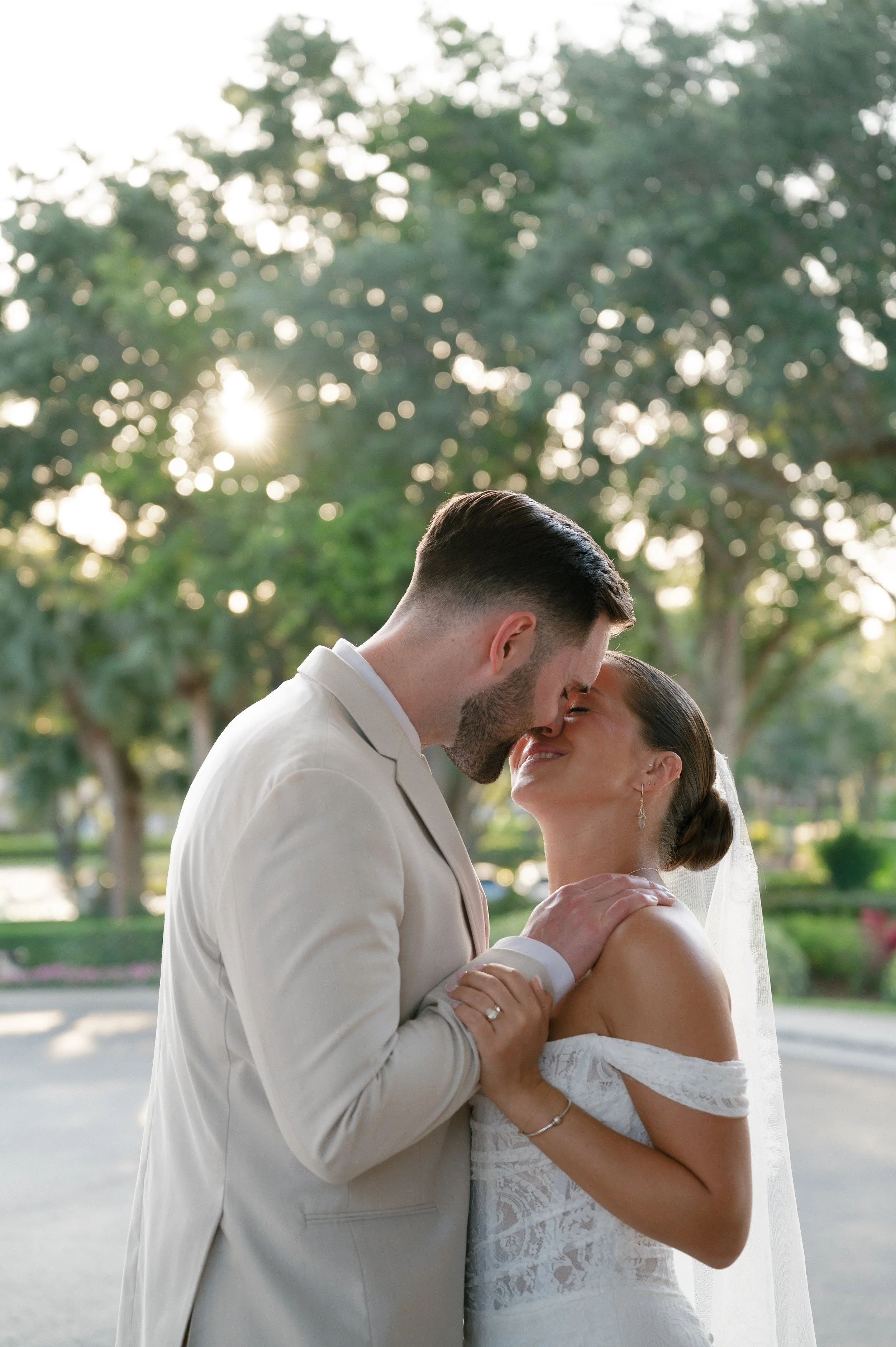 A bride and groom holding each other and about to kiss outdoors during the late afternoon or early evening with sunlight filtering through trees in the background.