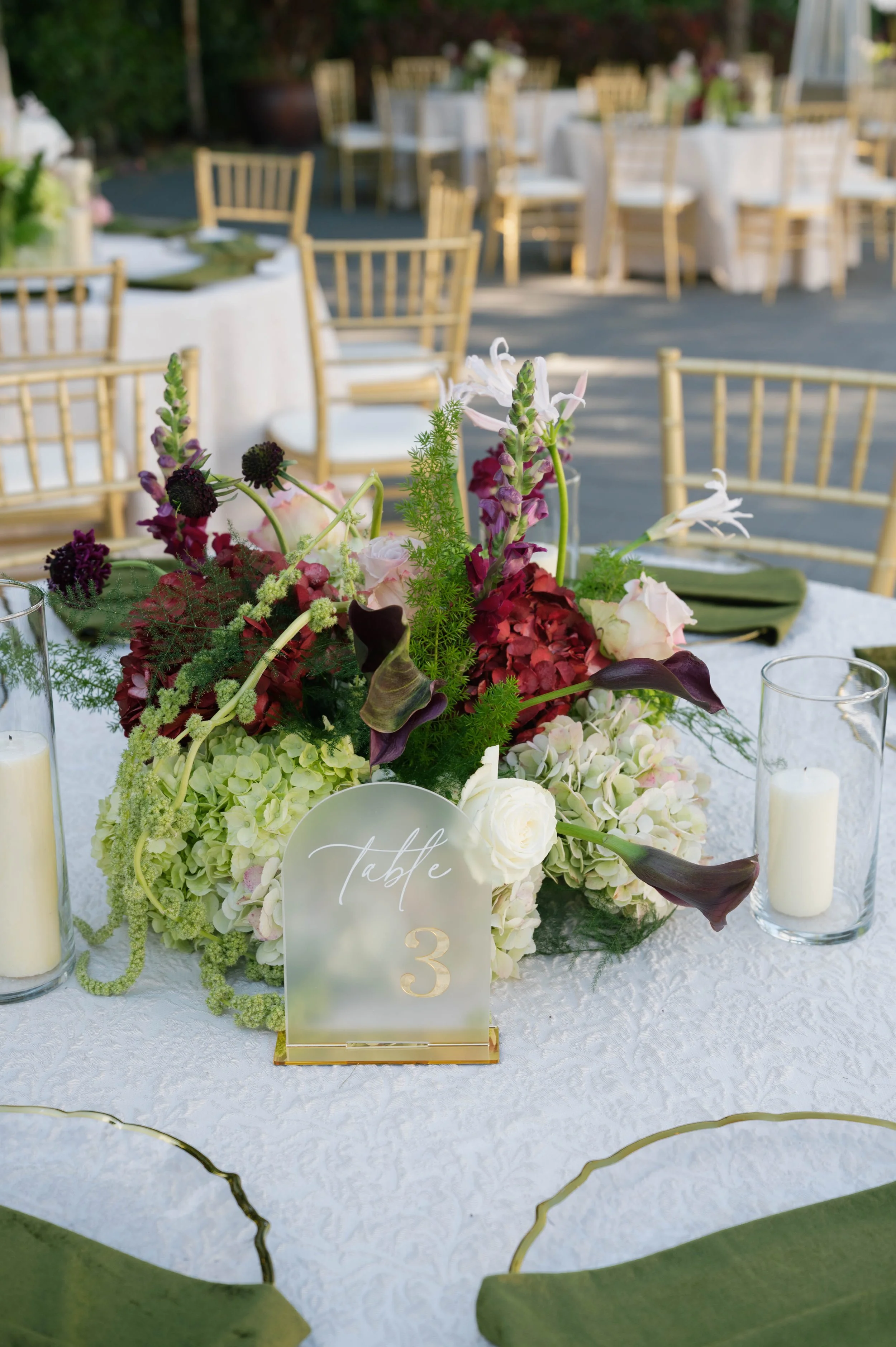 Close-up of a floral centerpiece on a white tablecloth, with a sign indicating it is table 3, surrounded by clear candle holders with white candles, at an outdoor event with gold chairs in the background.