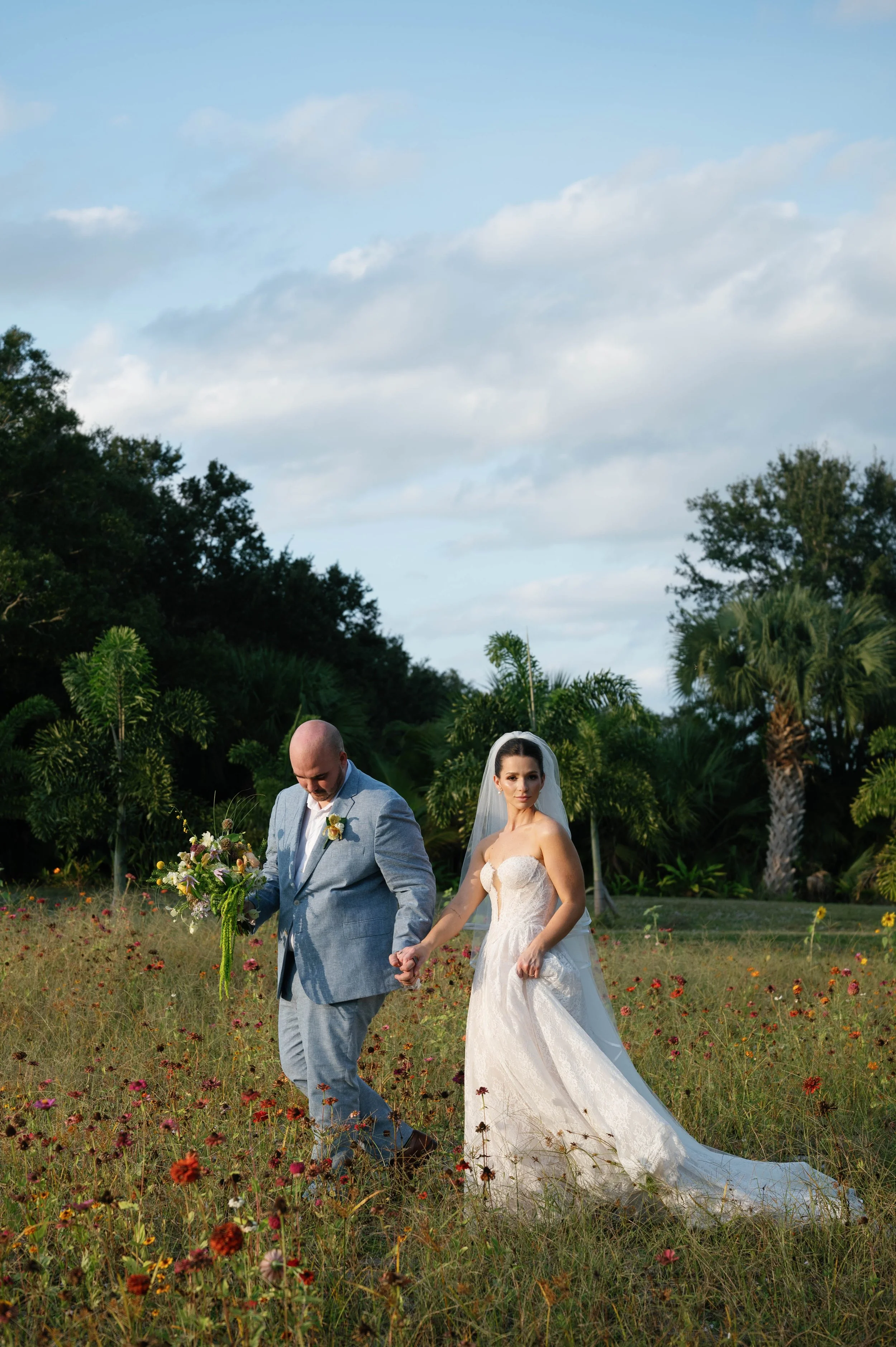 A bride and groom walk hand in hand through a field of wildflowers. The groom is looking down at the flowers, holding a bouquet, while the bride looks directly at the camera. The bride is wearing a white strapless wedding gown with a long train and a veil. The background features lush green trees, palm trees, and a partly cloudy sky.