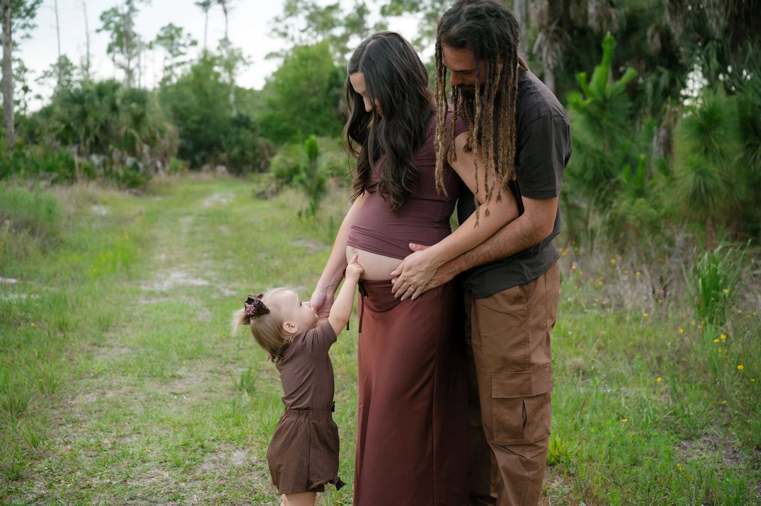 A pregnant woman, her partner, and a young girl outdoors in a green natural setting, with the girl touching the woman's belly and everyone smiling.