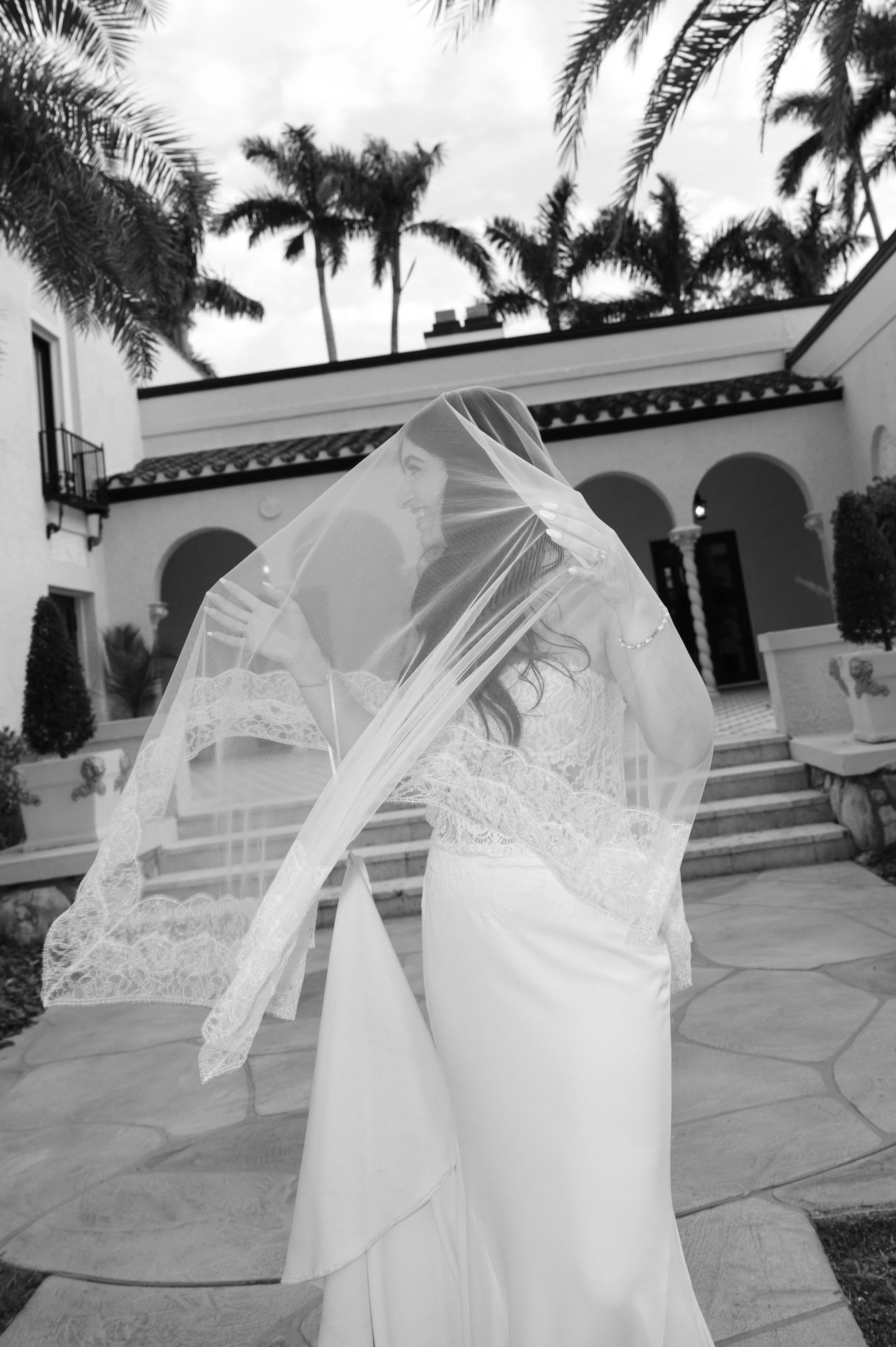 A bride in a wedding dress holding a lace veil over her face outdoors, with palm trees and a building in the background.
