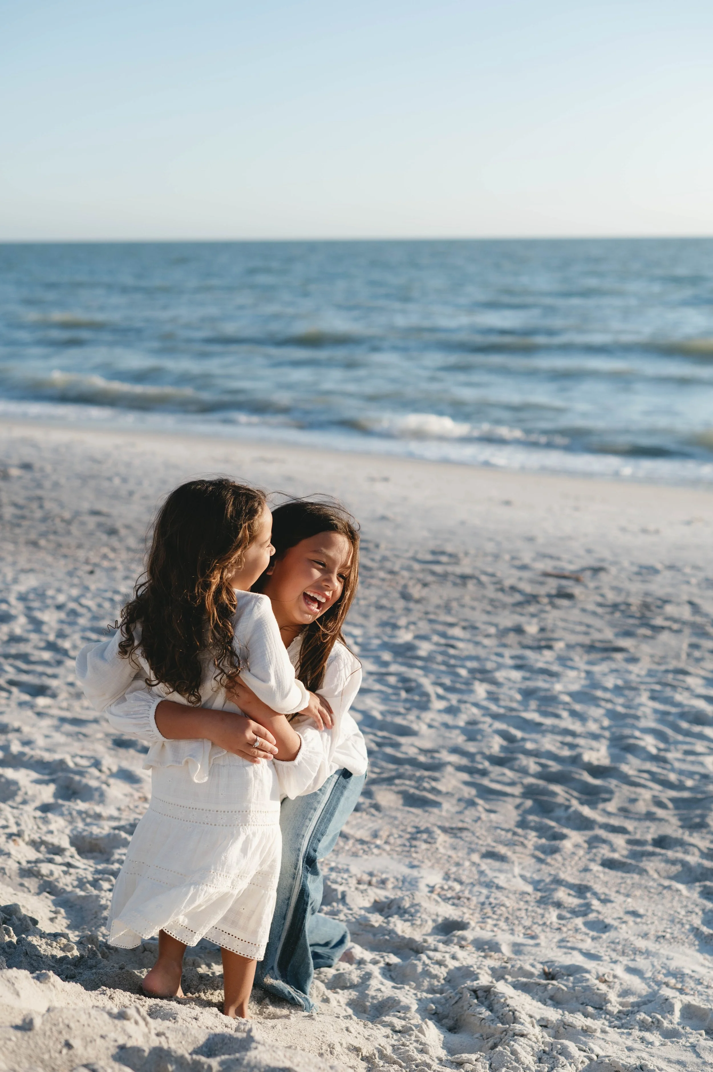 A woman and two young girls are on a sandy beach, smiling and laughing as they walk. The woman is carrying one girl in her arms, while the second girl walks close by. The ocean waves are gently rolling in the background under a clear sky.