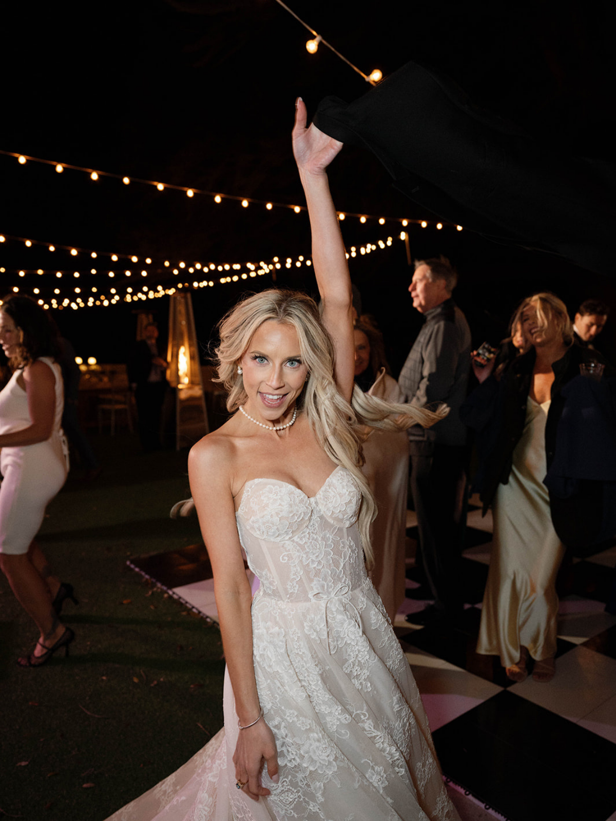 A woman in a wedding dress dancing at a celebration under string lights at night.