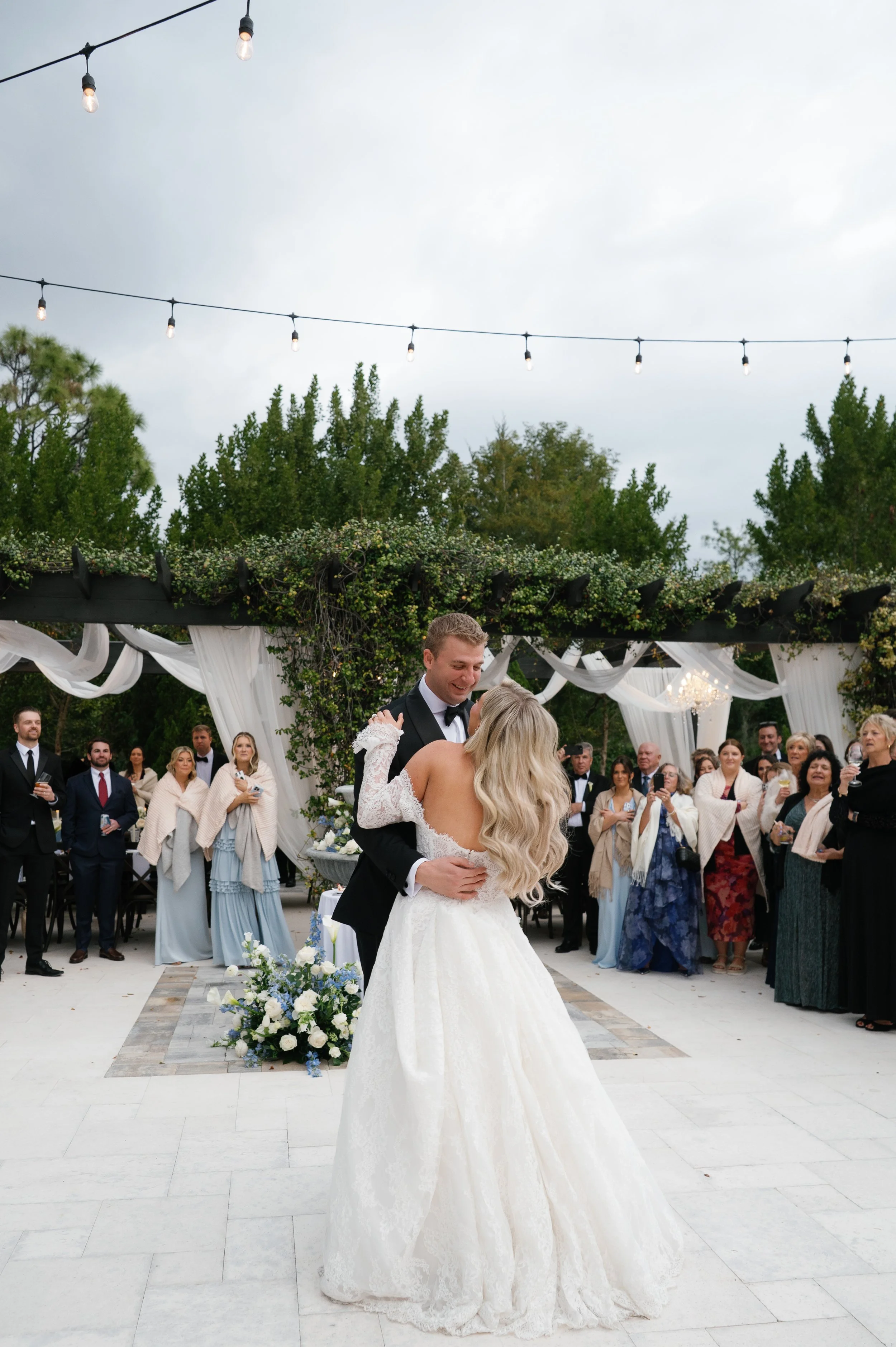 A bride and groom dance at their outdoor wedding ceremony, surrounded by guests, with string lights overhead and trees in the background.