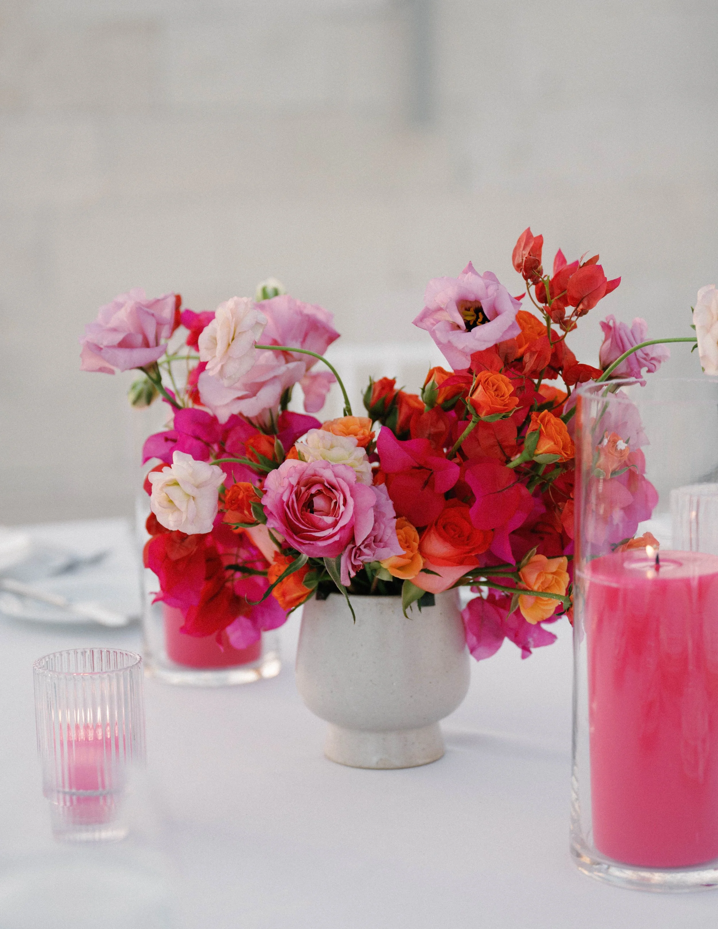 A white vase holding a colorful bouquet of pink, red, and orange flowers, placed on a white table, with pink candles and empty glasses around.