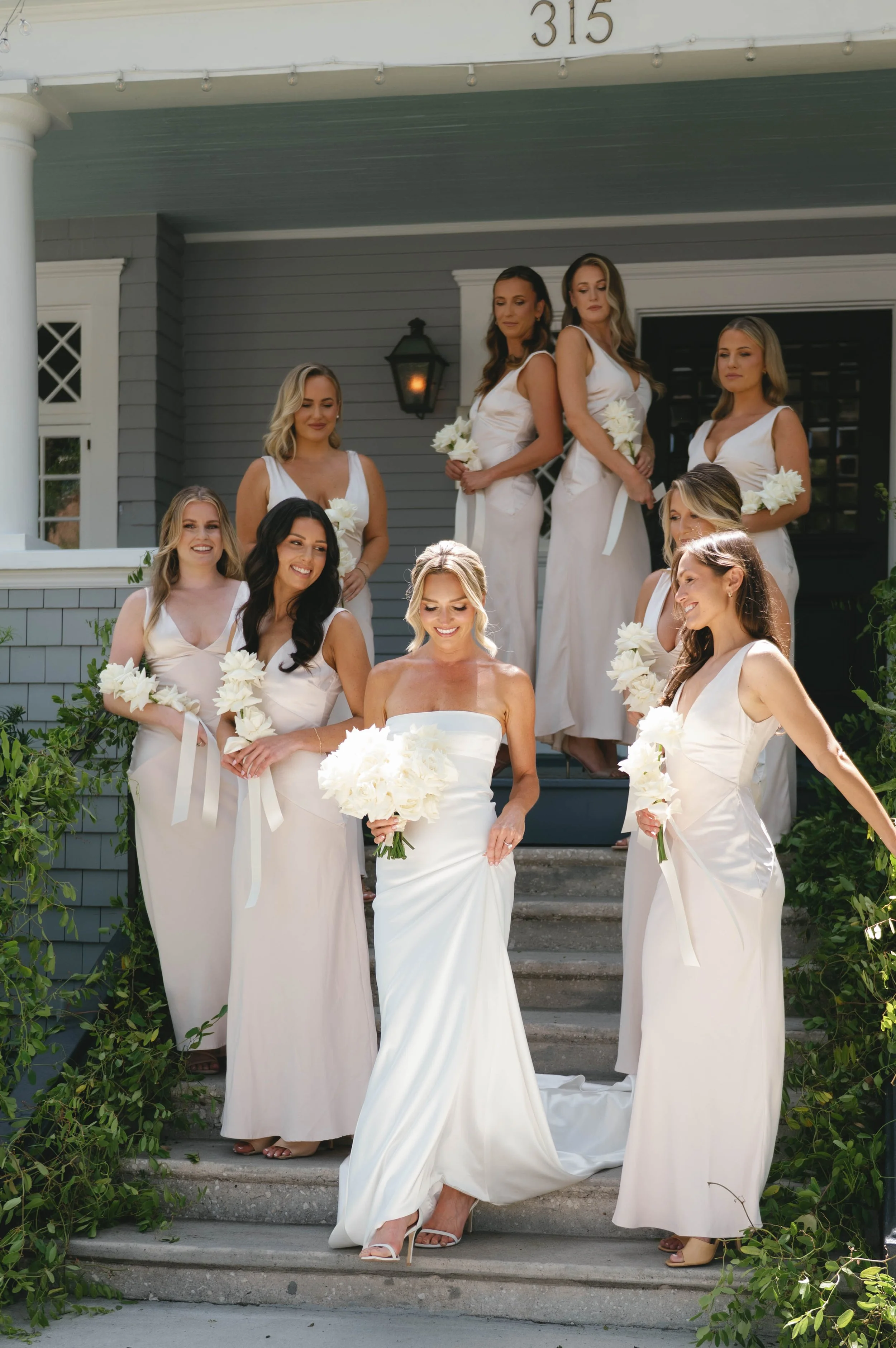 A bride and her bridesmaids standing on the steps outside a house, all wearing long cream-colored dresses and holding white bouquets.