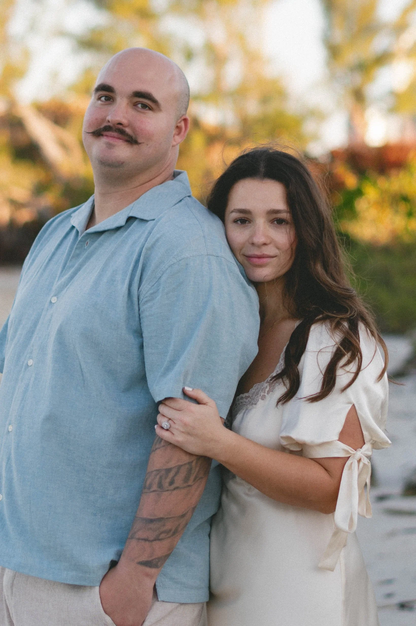 A man with a bald head and a mustache wearing a light blue shirt, standing outdoors with a woman with long dark hair and light skin, wearing a cream-colored dress with lace details, embracing each other with trees in the background during a sunny day
