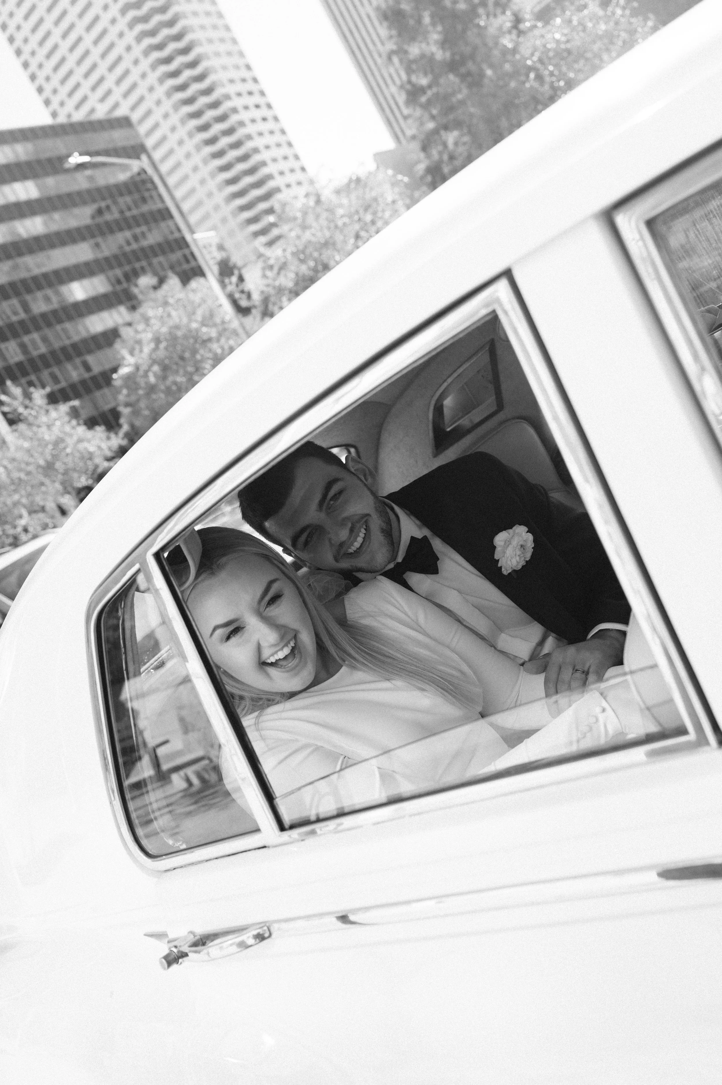 Black and white photo of a smiling bride and groom sitting in a vintage convertible car during their wedding, with tall modern buildings and trees in the background.