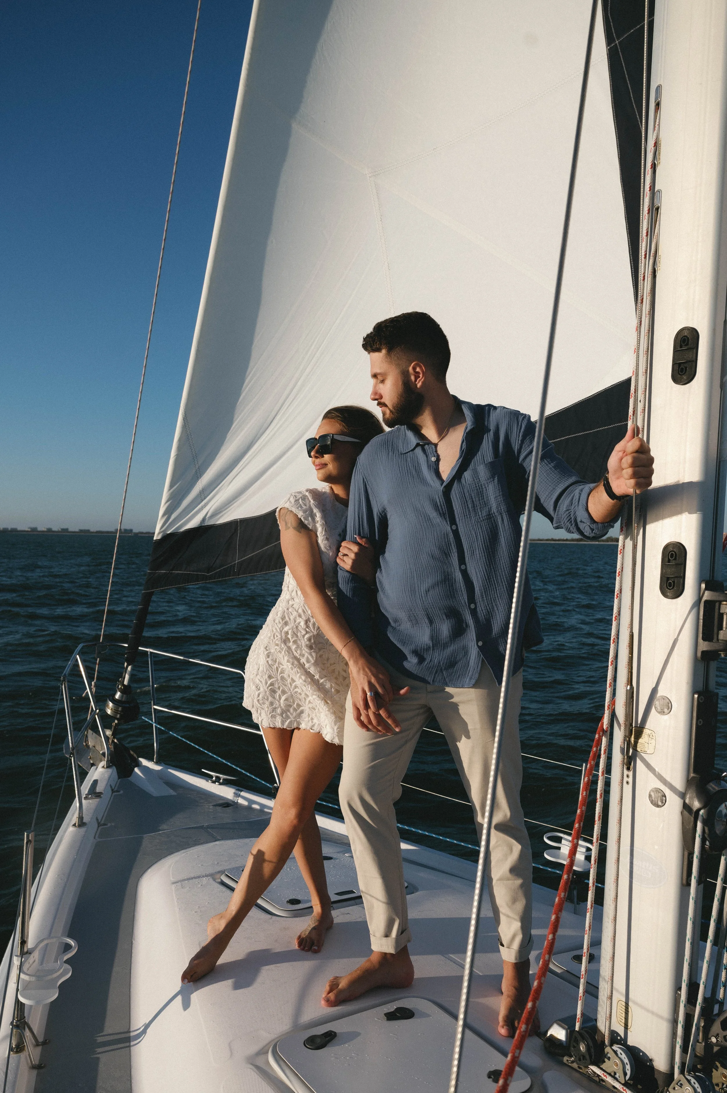 A couple standing close together on a sailboat, smiling, with the woman wearing sunglasses and a white lace dress, and the man wearing a blue shirt and beige pants, holding onto the boat's structure, on a sunny day with water in the background.