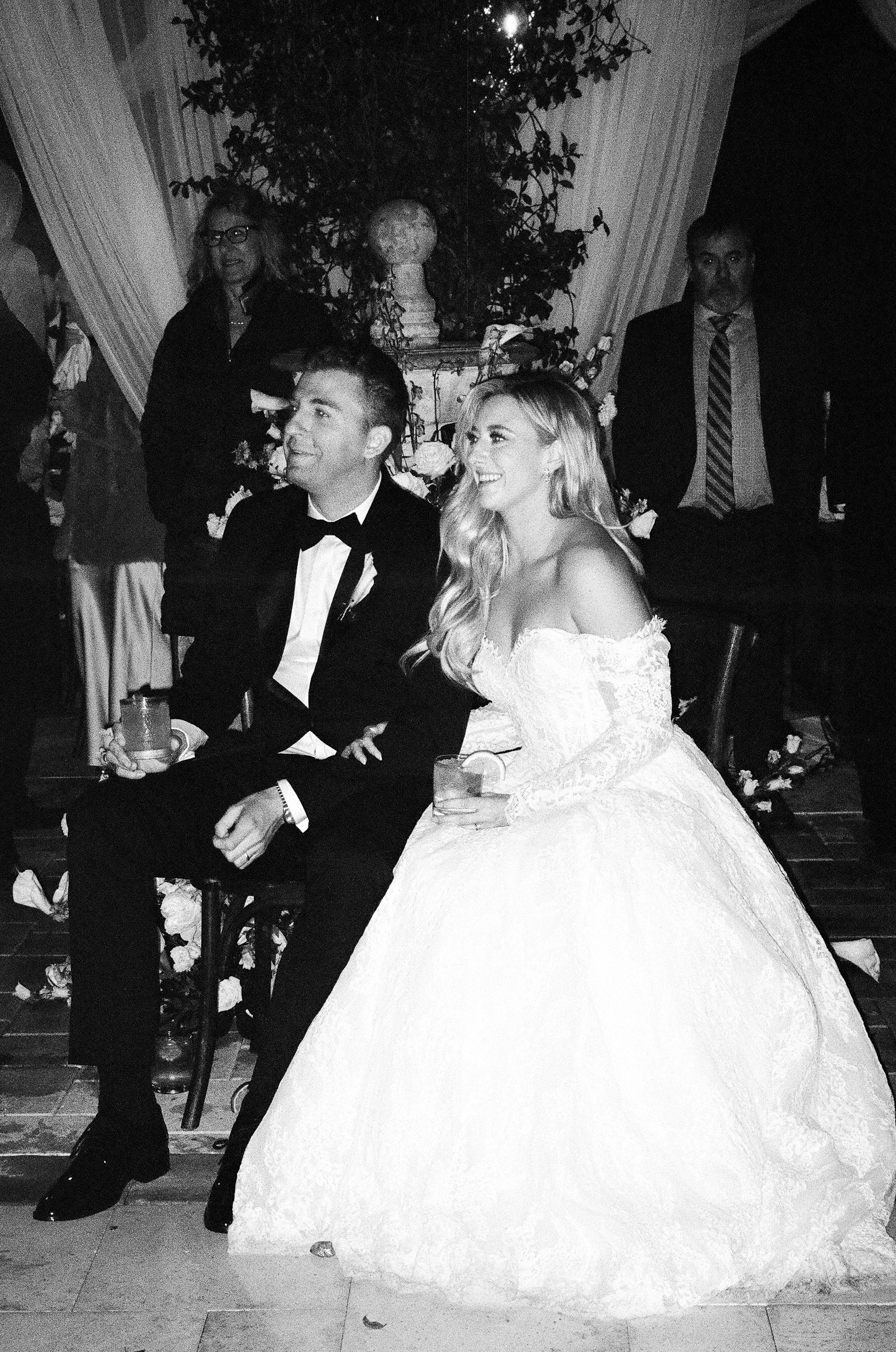 A black-and-white photo of a bride and groom seated at a wedding reception. The bride is wearing a strapless wedding gown with lace details and has long, wavy hair. The groom, wearing a tuxedo with a bow tie, is smiling. There are two guests standing