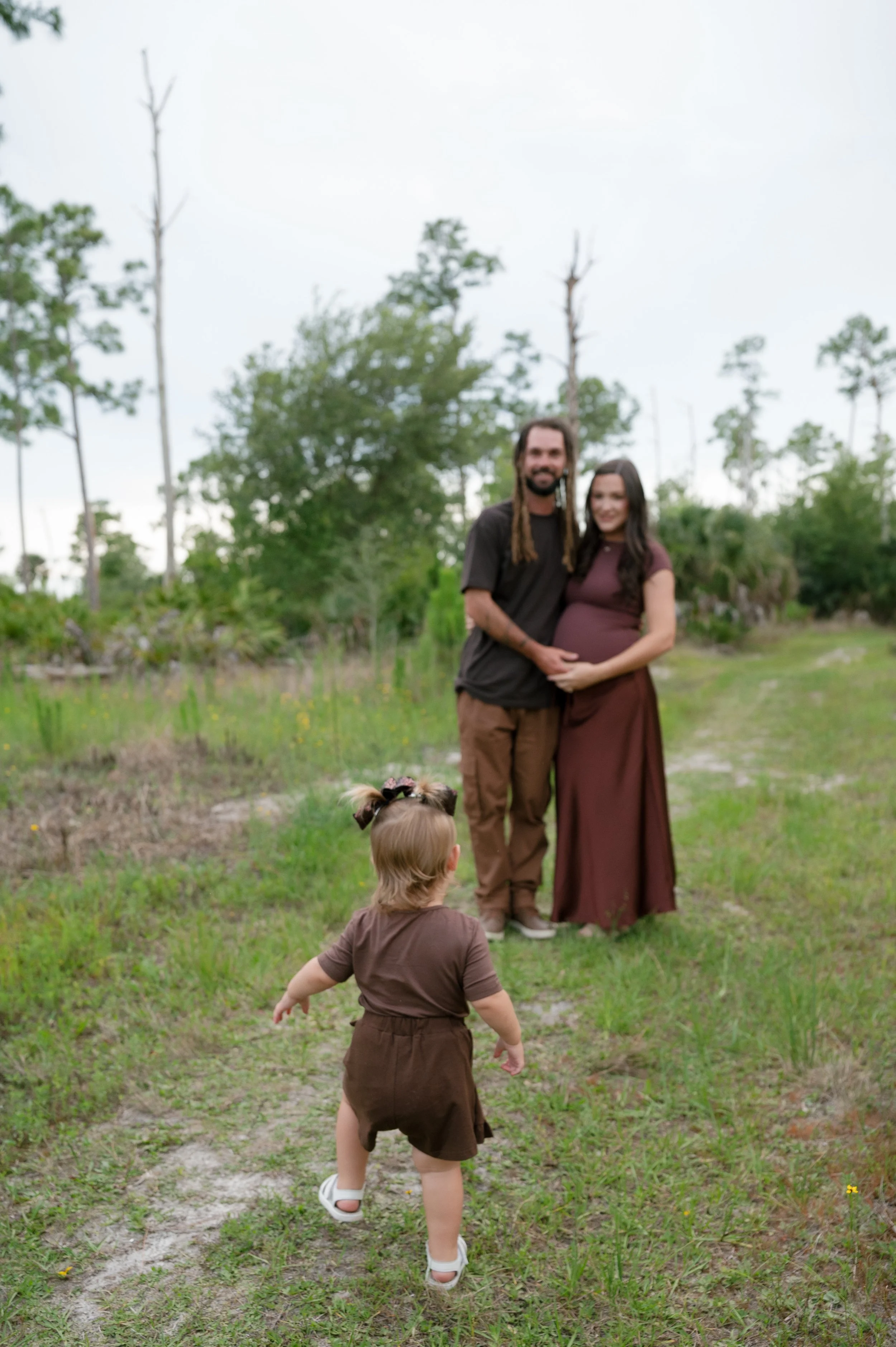 A family outdoors in a grassy area with trees; a man and woman stand together, with the woman showing a pregnant belly, and a young girl in the foreground walking towards them.