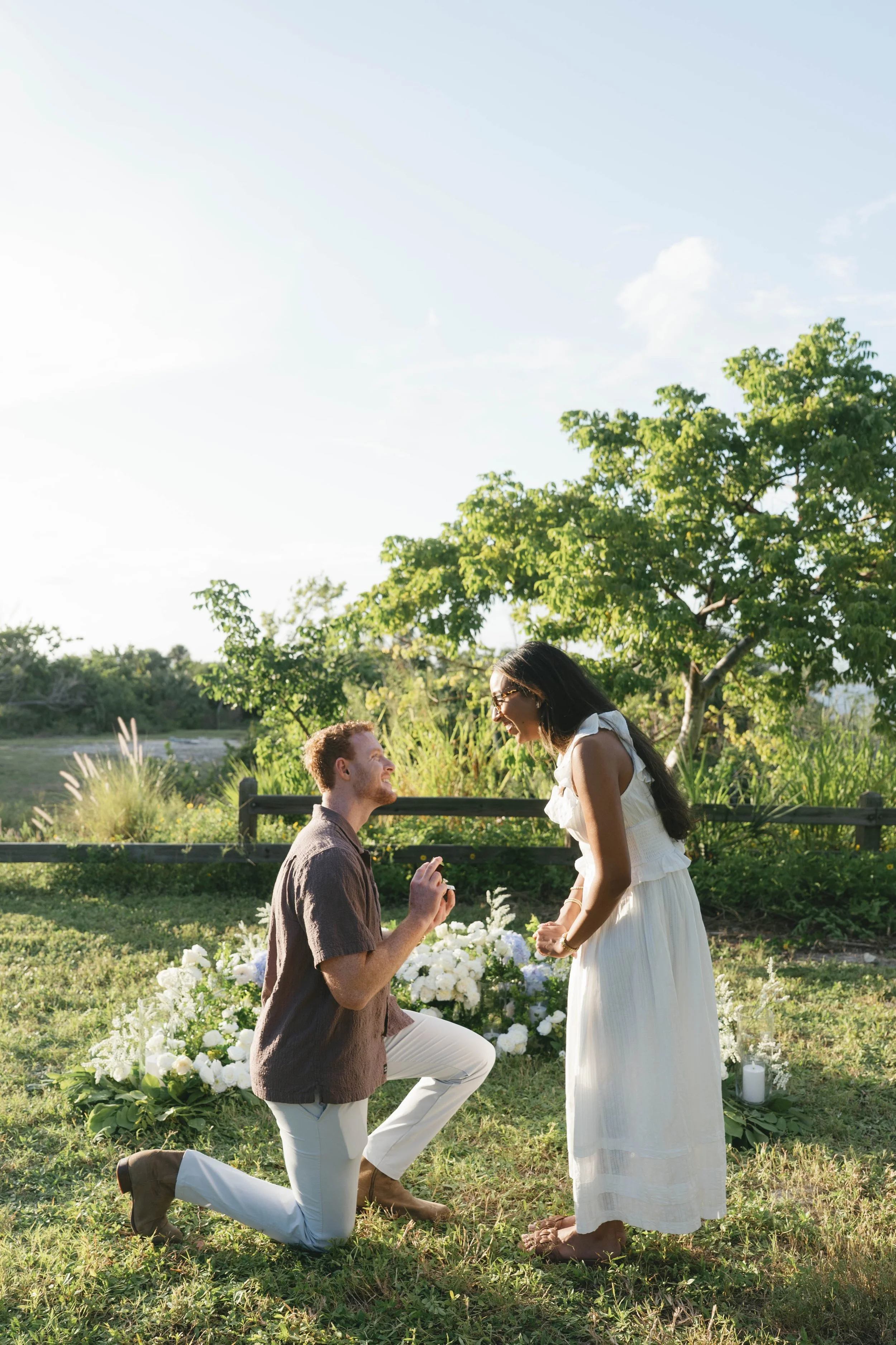 A man proposes marriage to a woman in a garden, with a man on one knee holding a ring, and the woman smiling and leaning towards him, surrounded by white flowers and lush greenery, during the daytime.