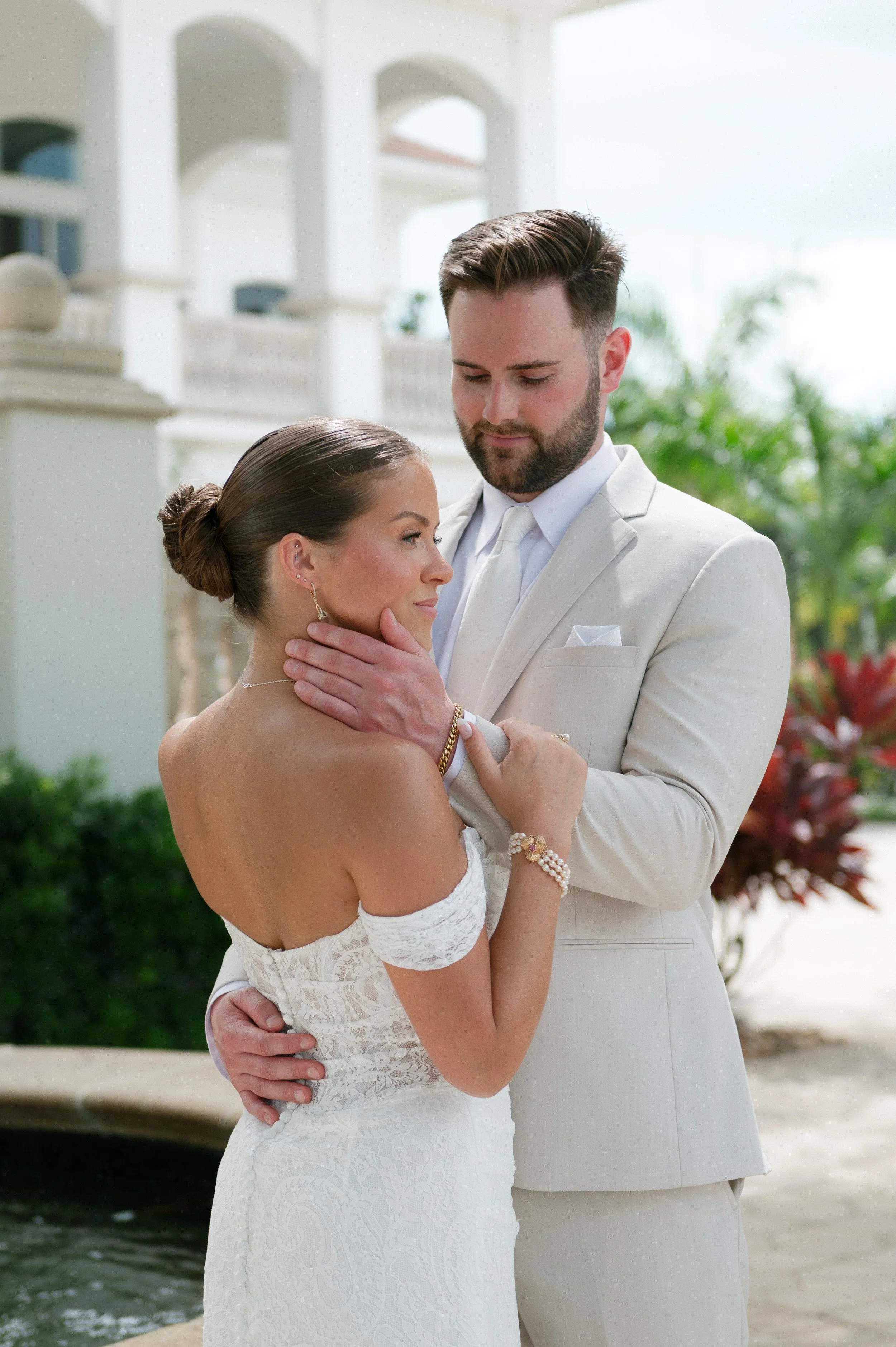 A bride and groom embrace outdoors near a body of water. The bride is in a white lace off-the-shoulder dress with her hair in a bun. The groom is in a light-colored suit with a white shirt and tie, with his hand gently touching her face and his other