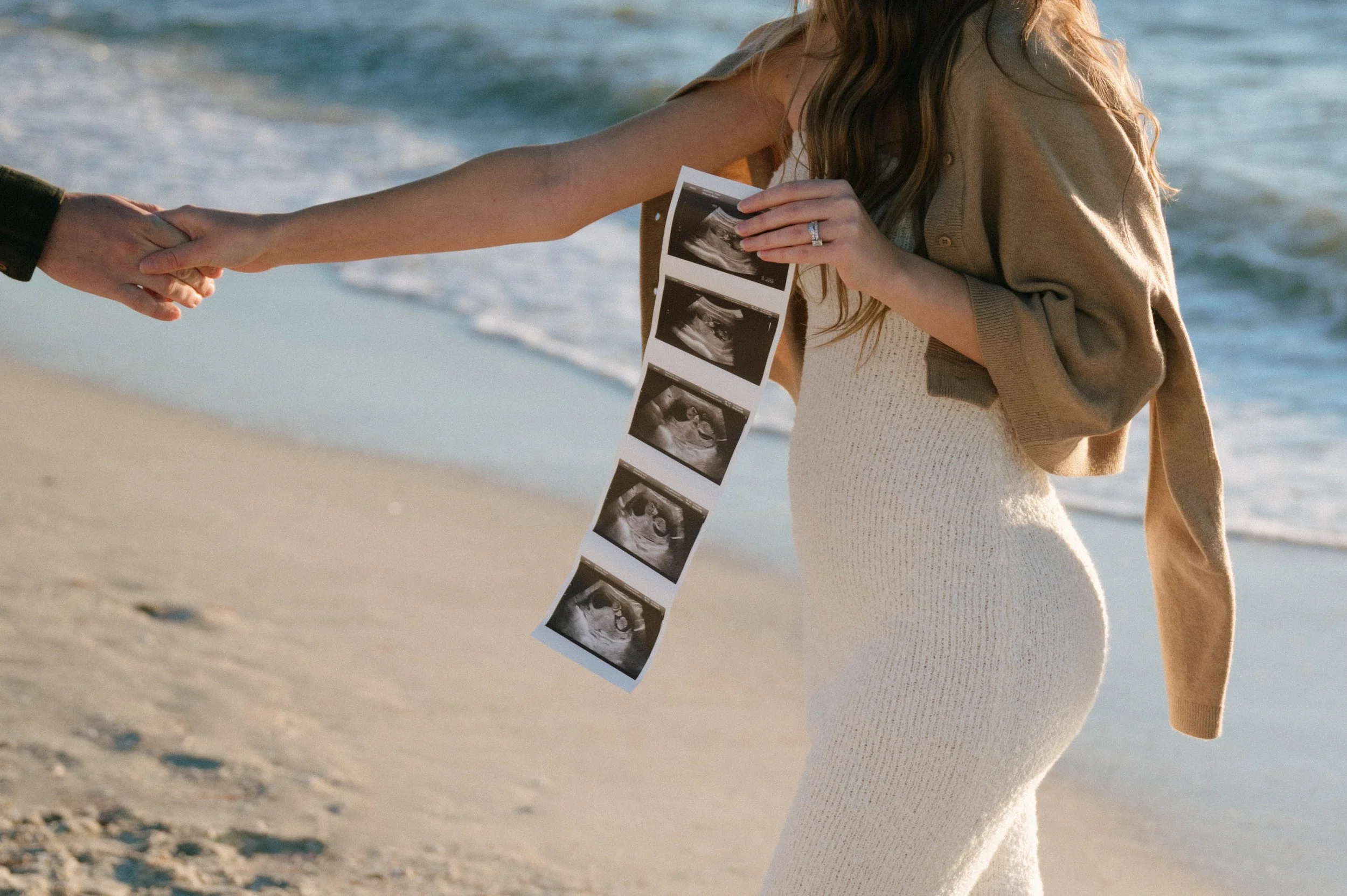 A woman at the beach holding ultrasound images of a fetus.