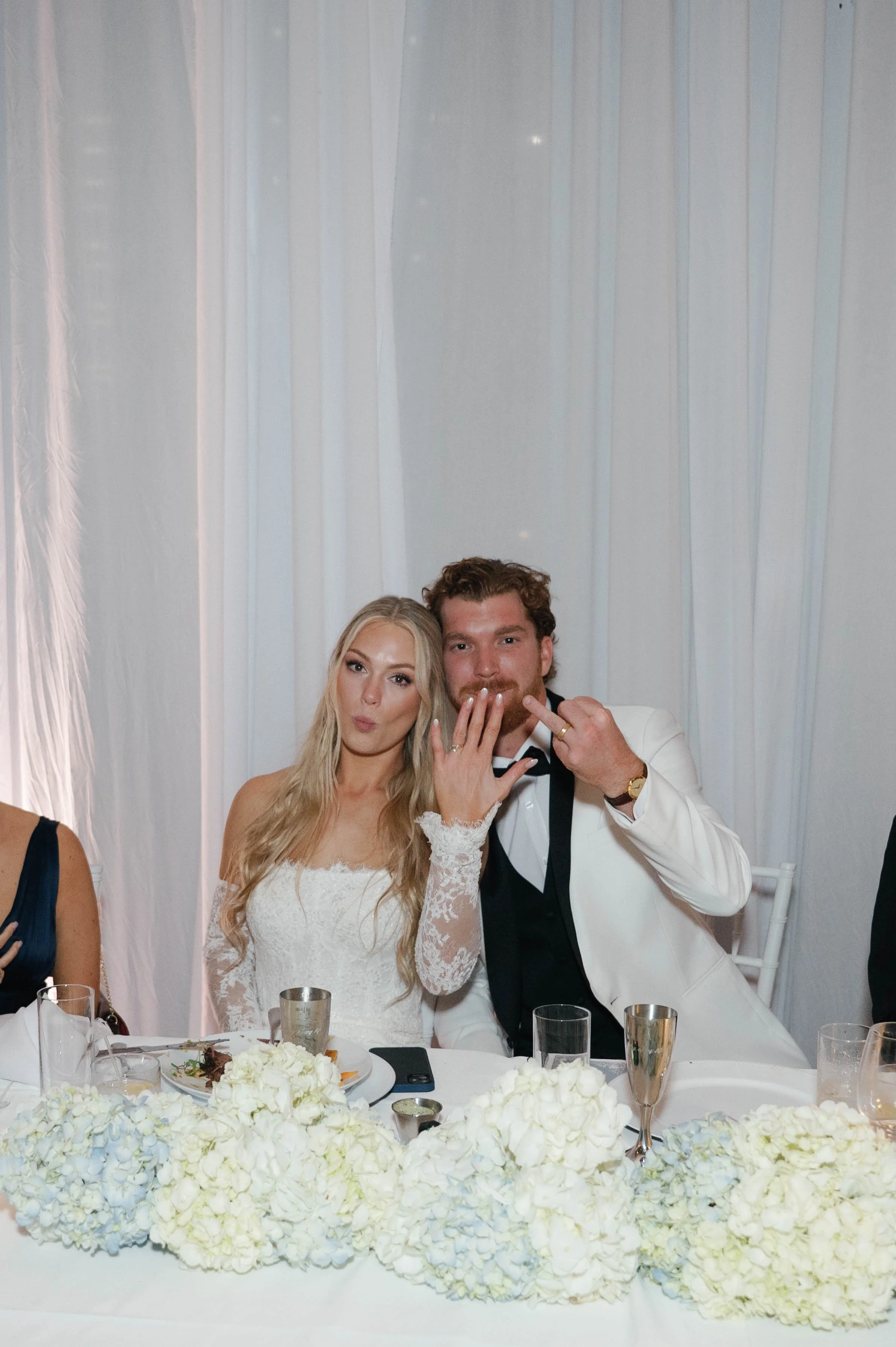 A newlywed couple at their wedding reception. The bride is wearing a white lace wedding dress and the groom is in a white tuxedo jacket with black bow tie. They are seated at a table decorated with white flowers, with plates of food and drinks in fro