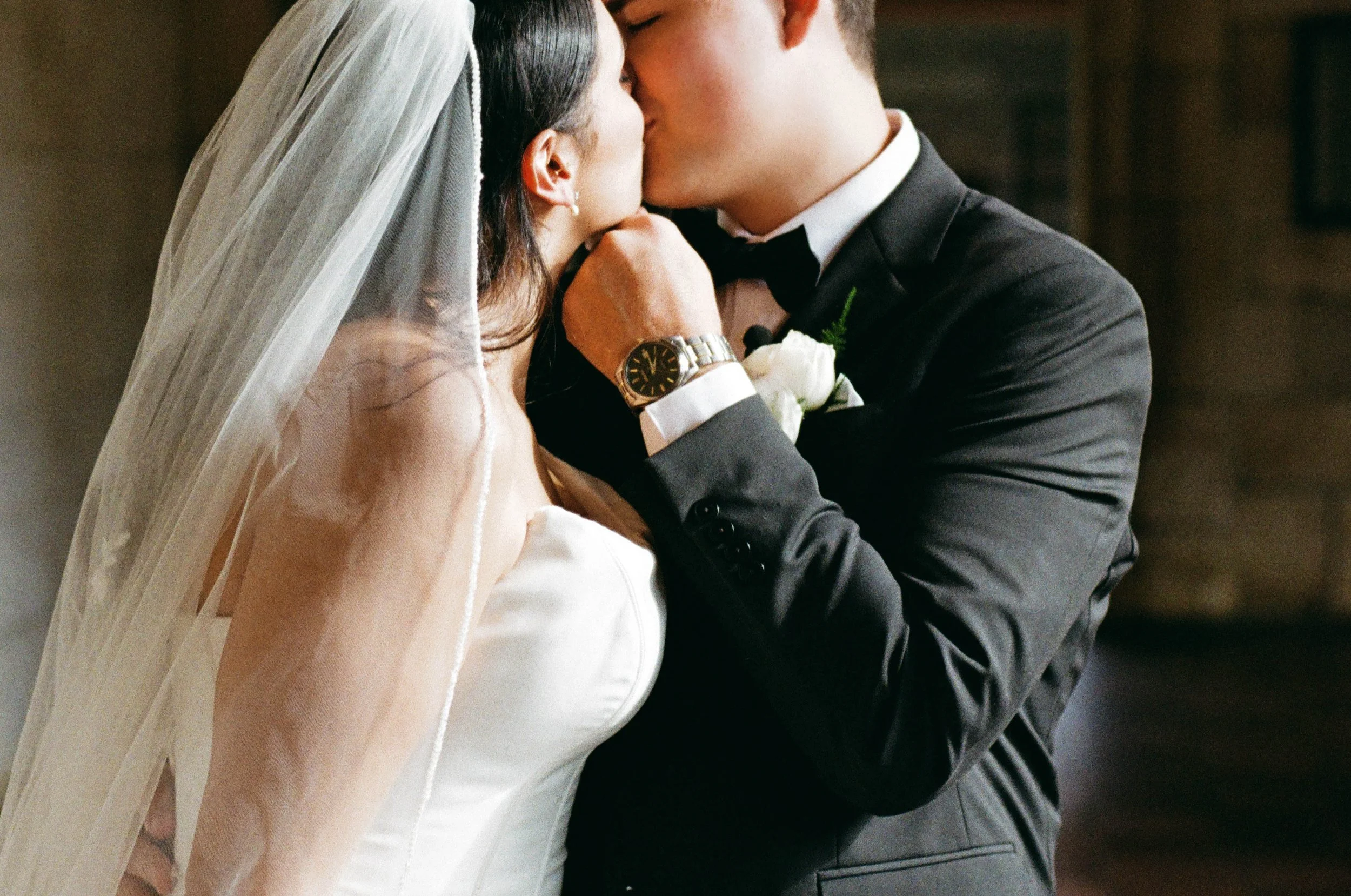 A bride and groom sharing a kiss, with the bride wearing a white wedding dress and veil, and the groom in a black tuxedo, both embracing closely.