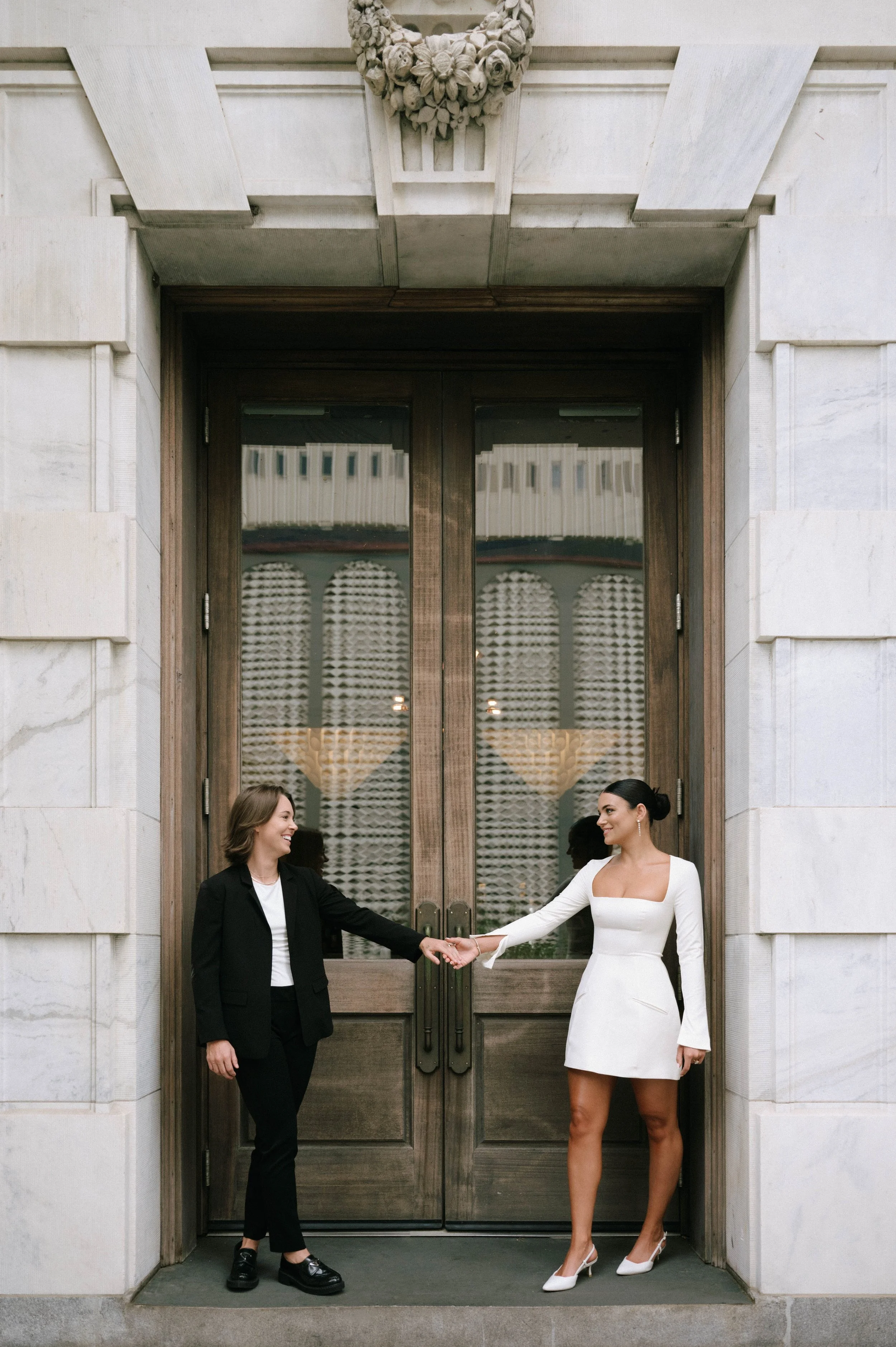 Two women in formal attire shake hands in front of a large wooden door with a floral wreath above it, inside a building with stone walls.