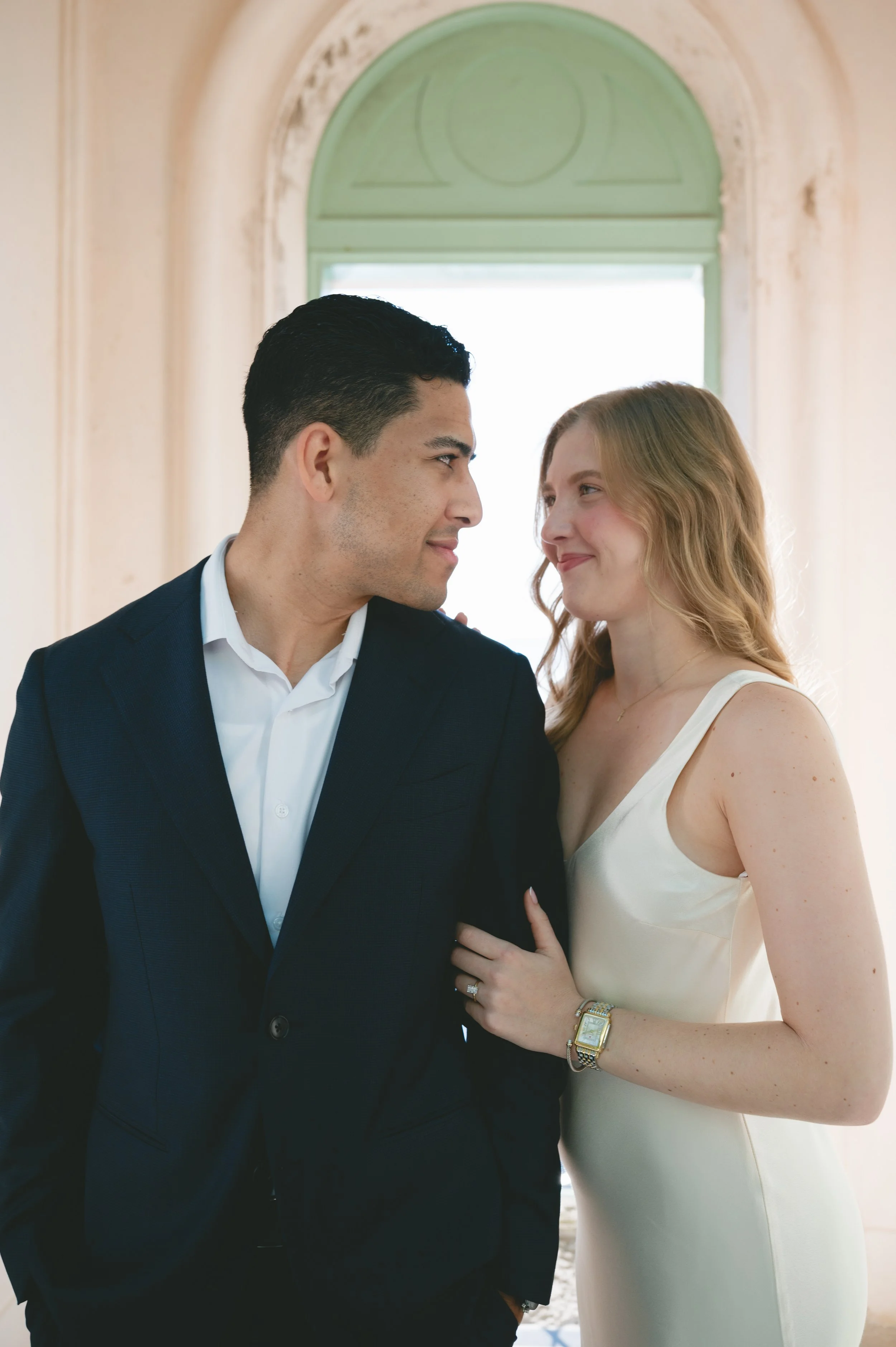 A man and woman are standing close together, facing each other, and smiling. The man is wearing a dark suit and white shirt, while the woman is in a white sleeveless dress, wearing a watch and a ring. They are indoors with a window and an arch in the