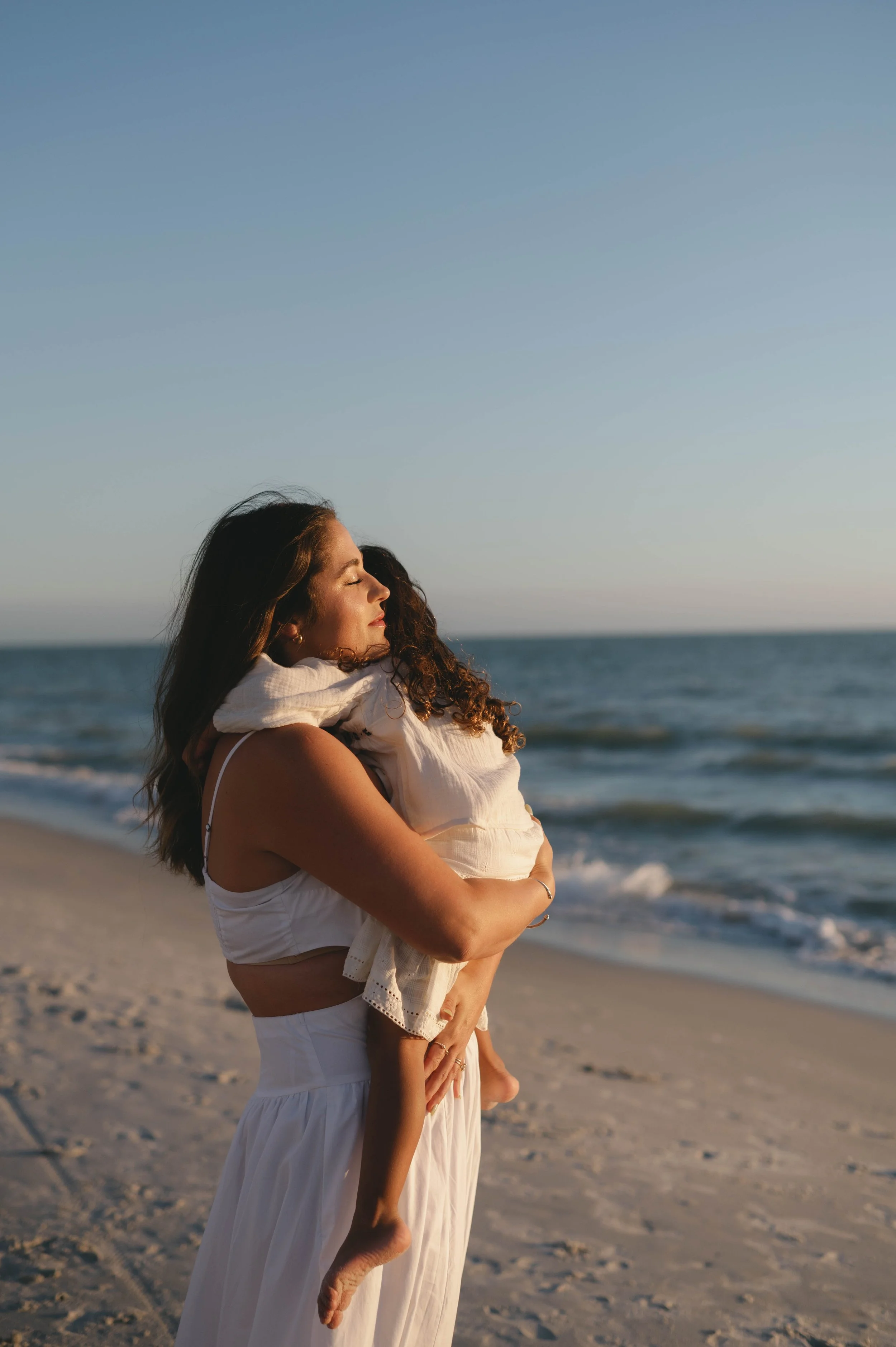 A woman is standing on a sandy beach, holding a young girl, both with eyes closed and enjoying the moment. The background features ocean waves and a clear sky.