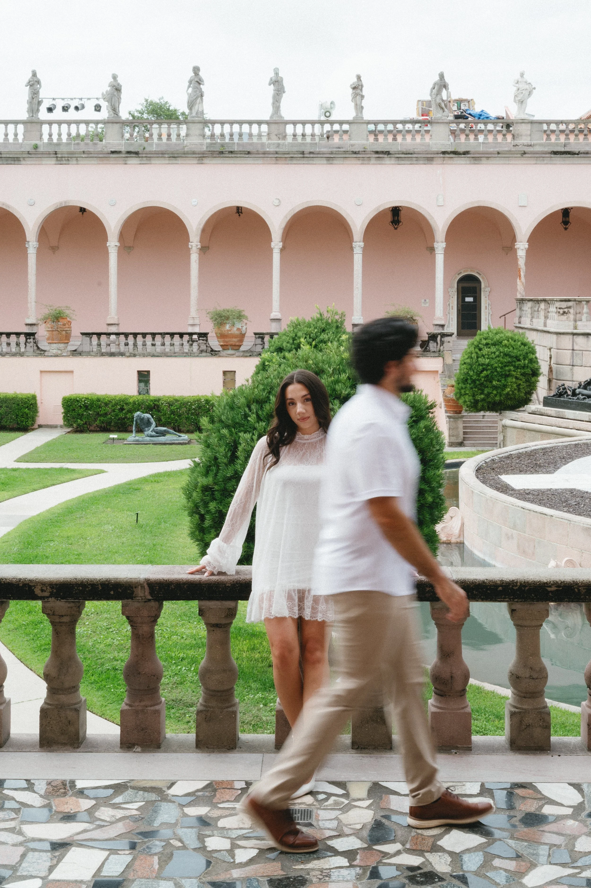 A woman in a white lace dress leaning on a stone railing in a garden with a man walking by in beige pants and brown shoes. In the background, there is a pink building with archways, potted plants, statues on the roof, and a lion sculpture on the lawn
