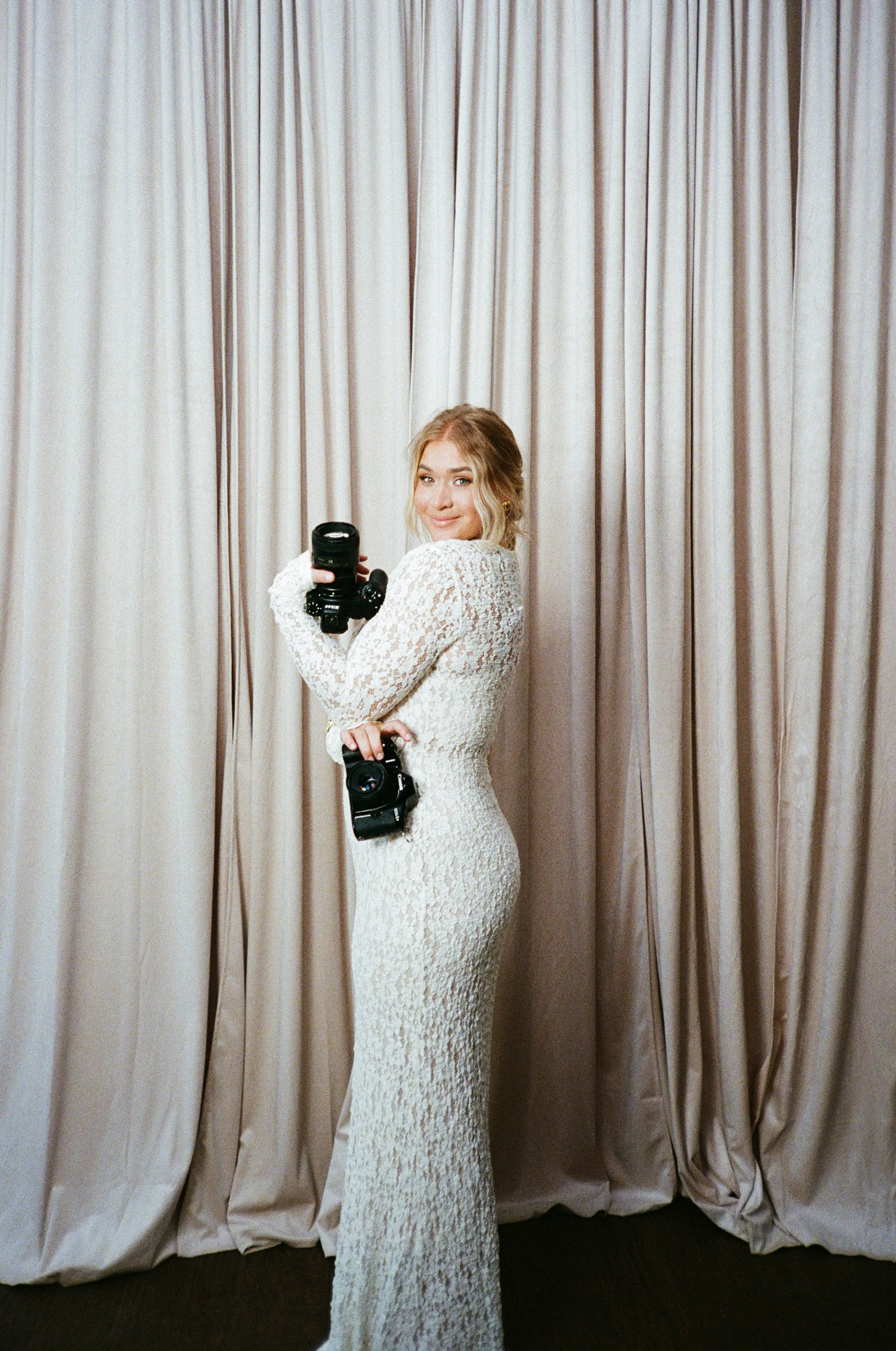 A woman in a long white lace dress holds a camera with a large lens, standing in front of beige curtains, smiling at the camera.