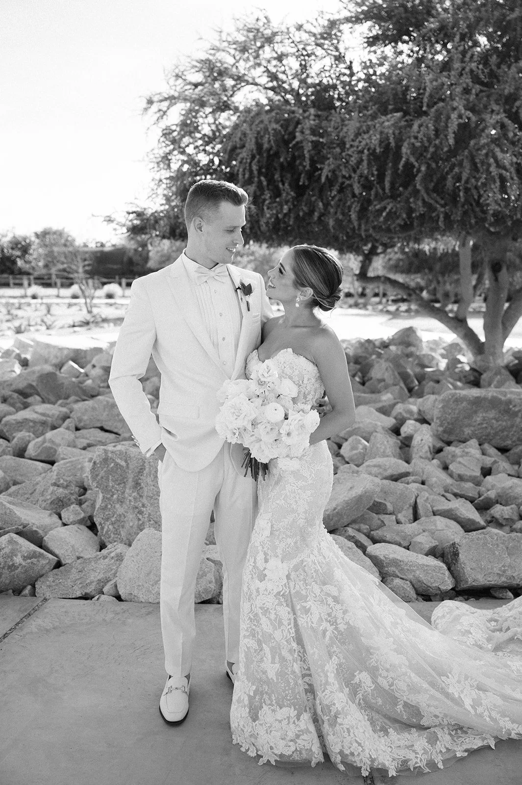 A black and white photo of a bride and groom standing outdoors on a rocky surface, with trees in the background. The bride is holding a large bouquet of flowers and wearing a strapless lace wedding gown. The groom is dressed in a white suit with a bo