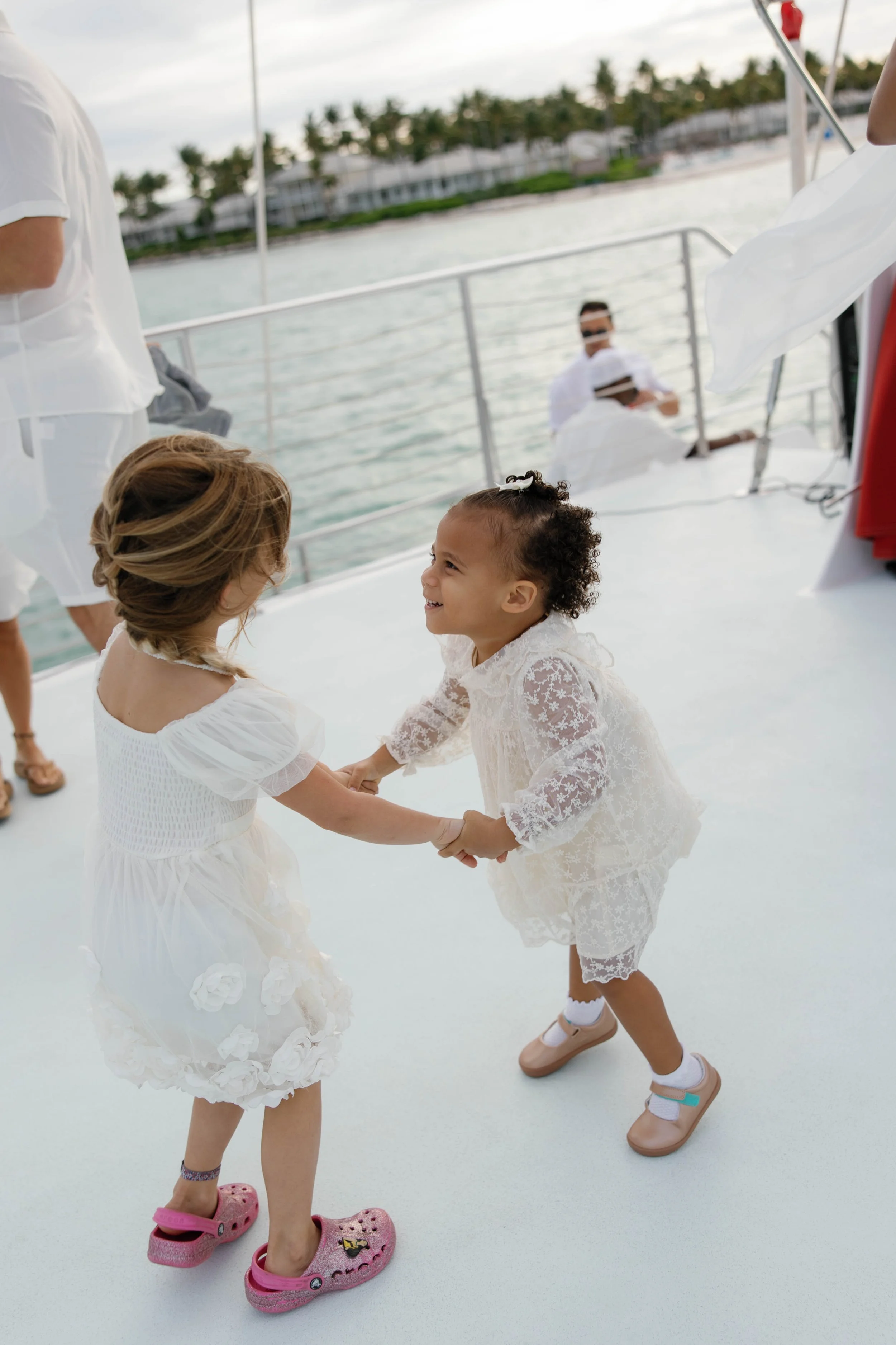 Two young girls in white lace dresses hold hands and dance on a white boat deck, barefoot and wearing casual shoes, while adults watch in the background.