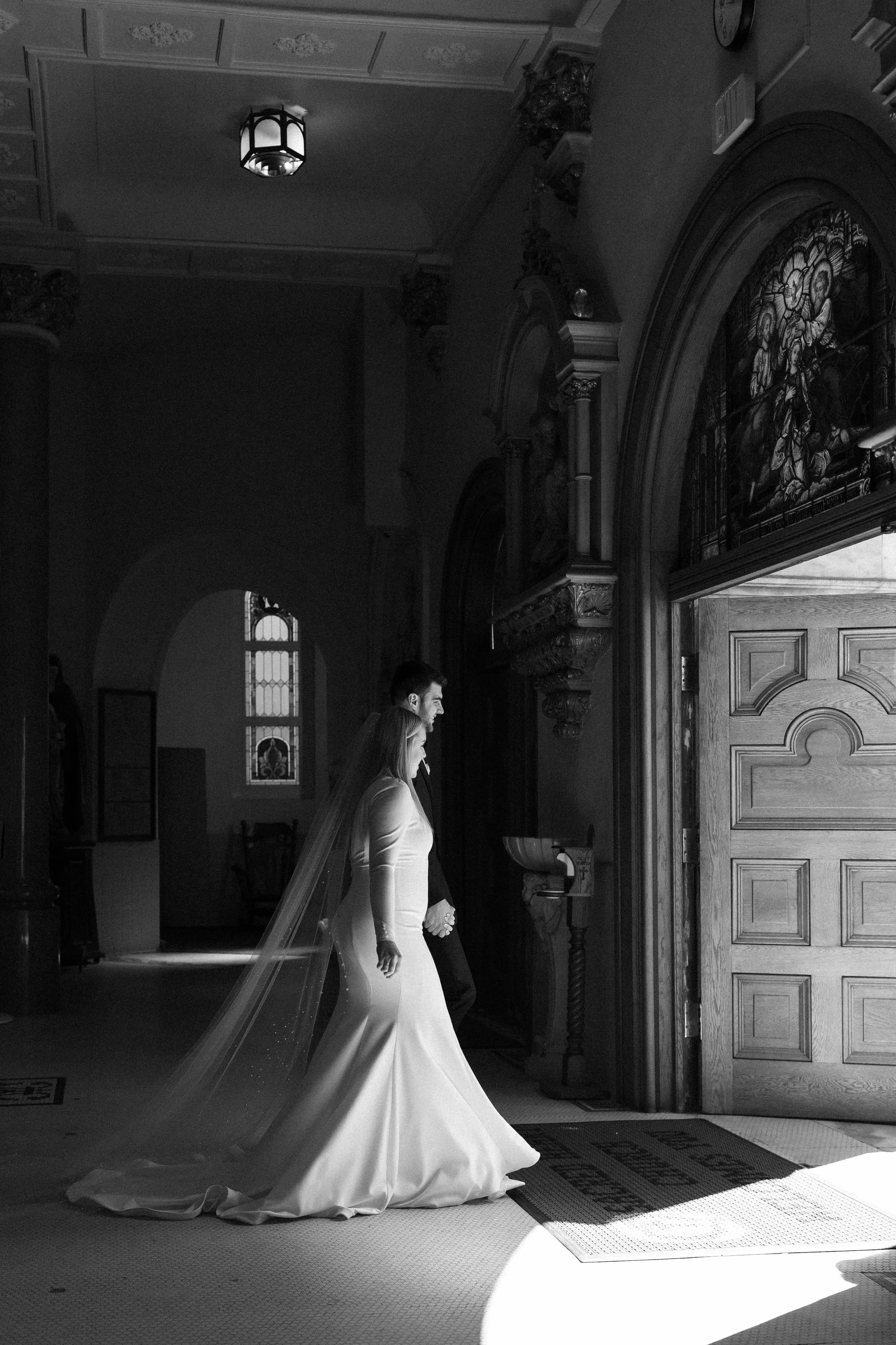 Bride and groom walking into a church for their wedding ceremony, with stained glass windows and ornate woodwork.