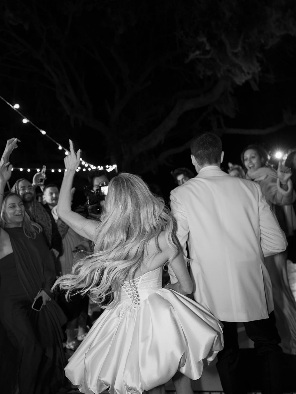 A black and white photo of a lively outdoor wedding reception at night, with guests dancing and celebrating around a bride and groom. String lights are overhead and large trees are in the background.