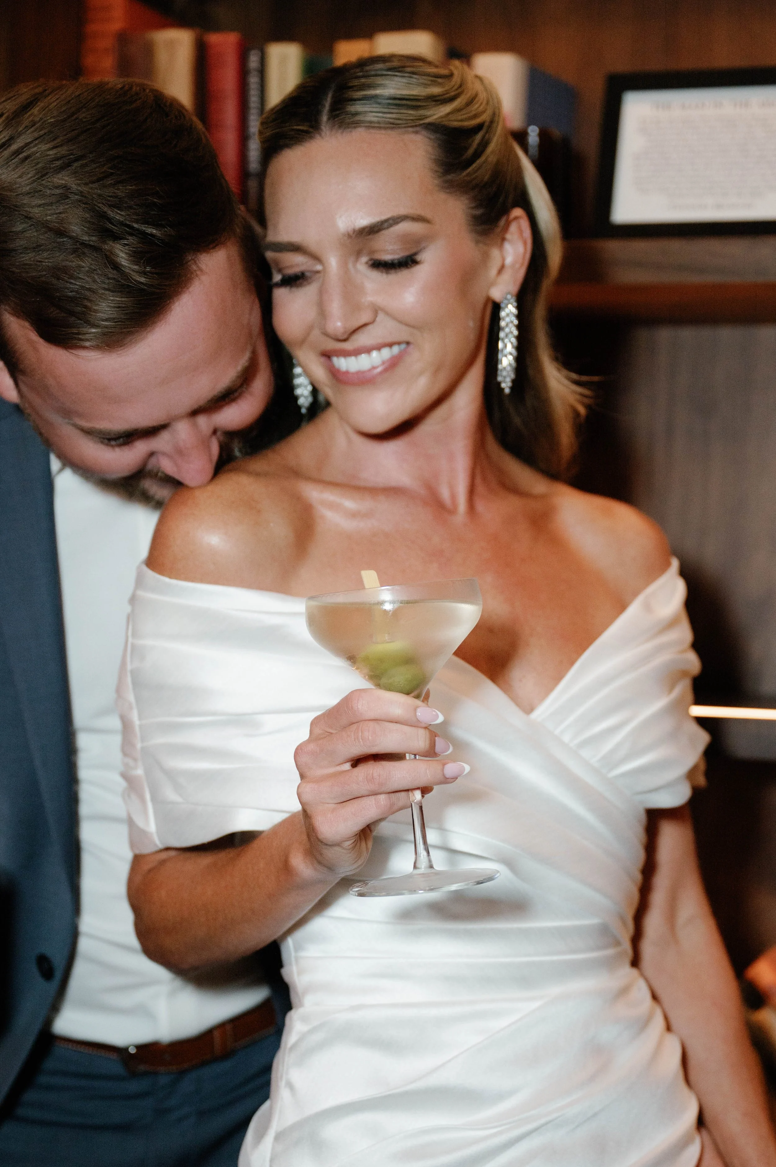 A woman in an off-shoulder white satin dress holding a champagne glass with green olives, smiling and leaning her head to a man in a dark suit.