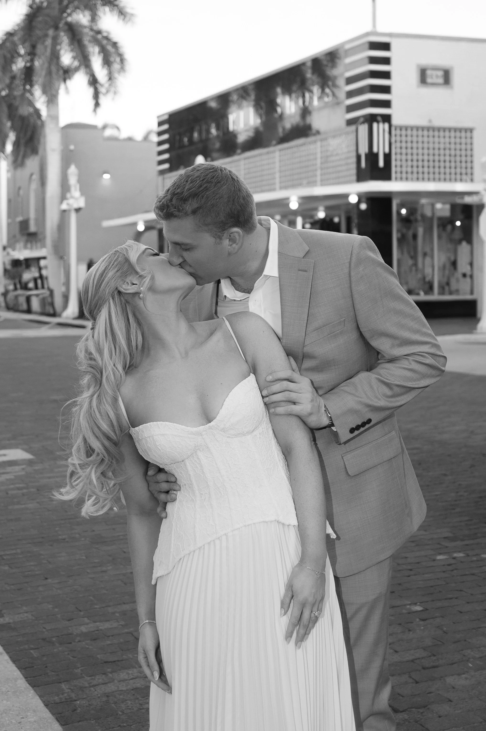 A couple dancing and kissing in a black-and-white photo outdoors, with a modern building and palm trees in the background.
