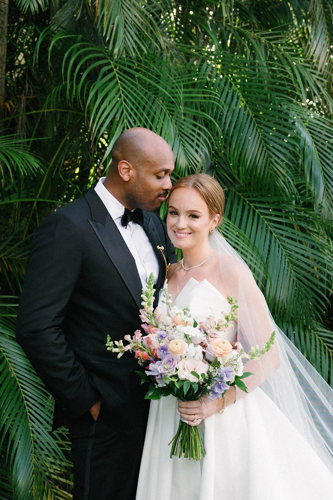 A bride and groom pose together for their wedding photo in front of lush green tropical plants. The groom is in a black tuxedo with a bow tie, and the bride is in a white wedding gown holding a bouquet of pastel-colored flowers.