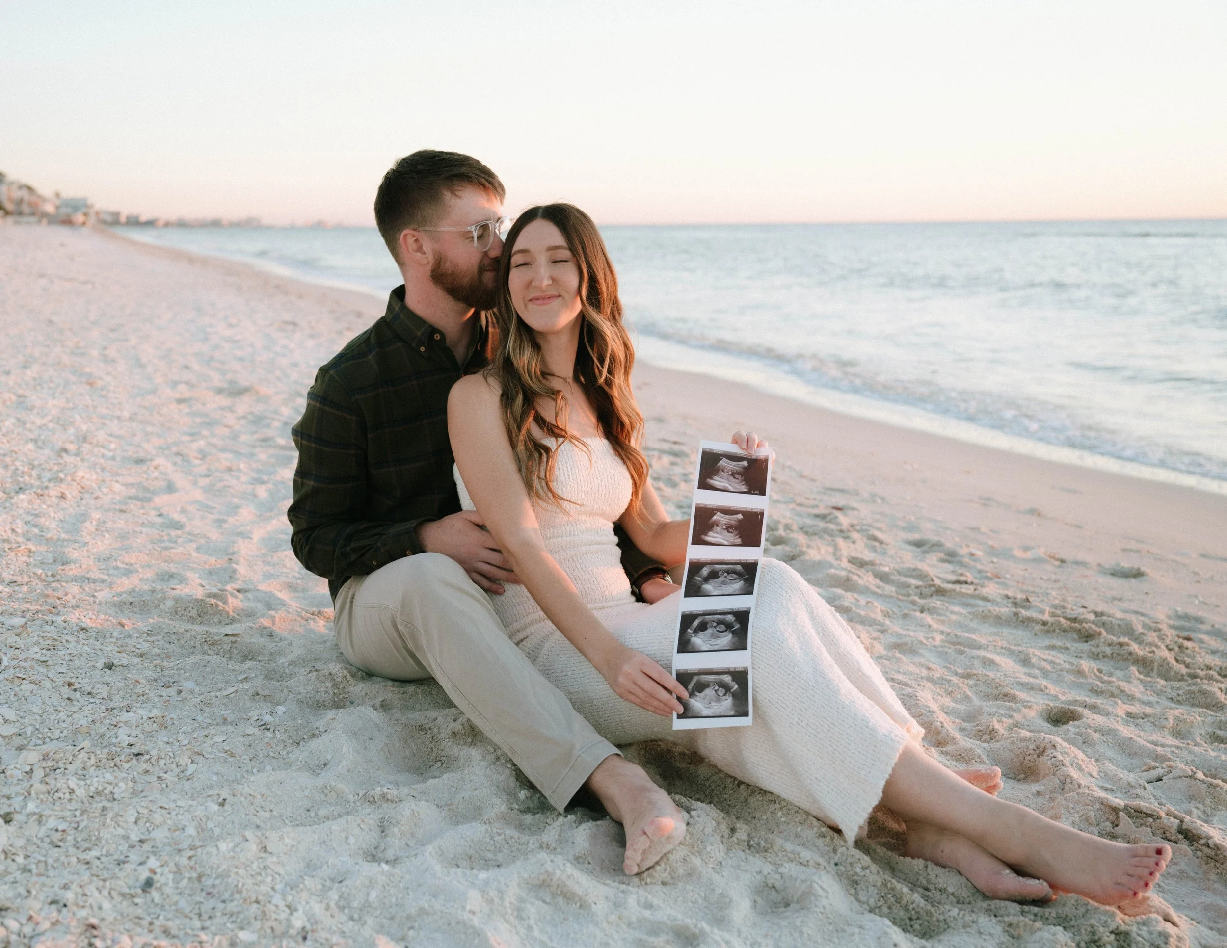 A couple sitting on a sandy beach, with the woman holding ultrasound images, as the man kisses her on the cheek, during sunset.