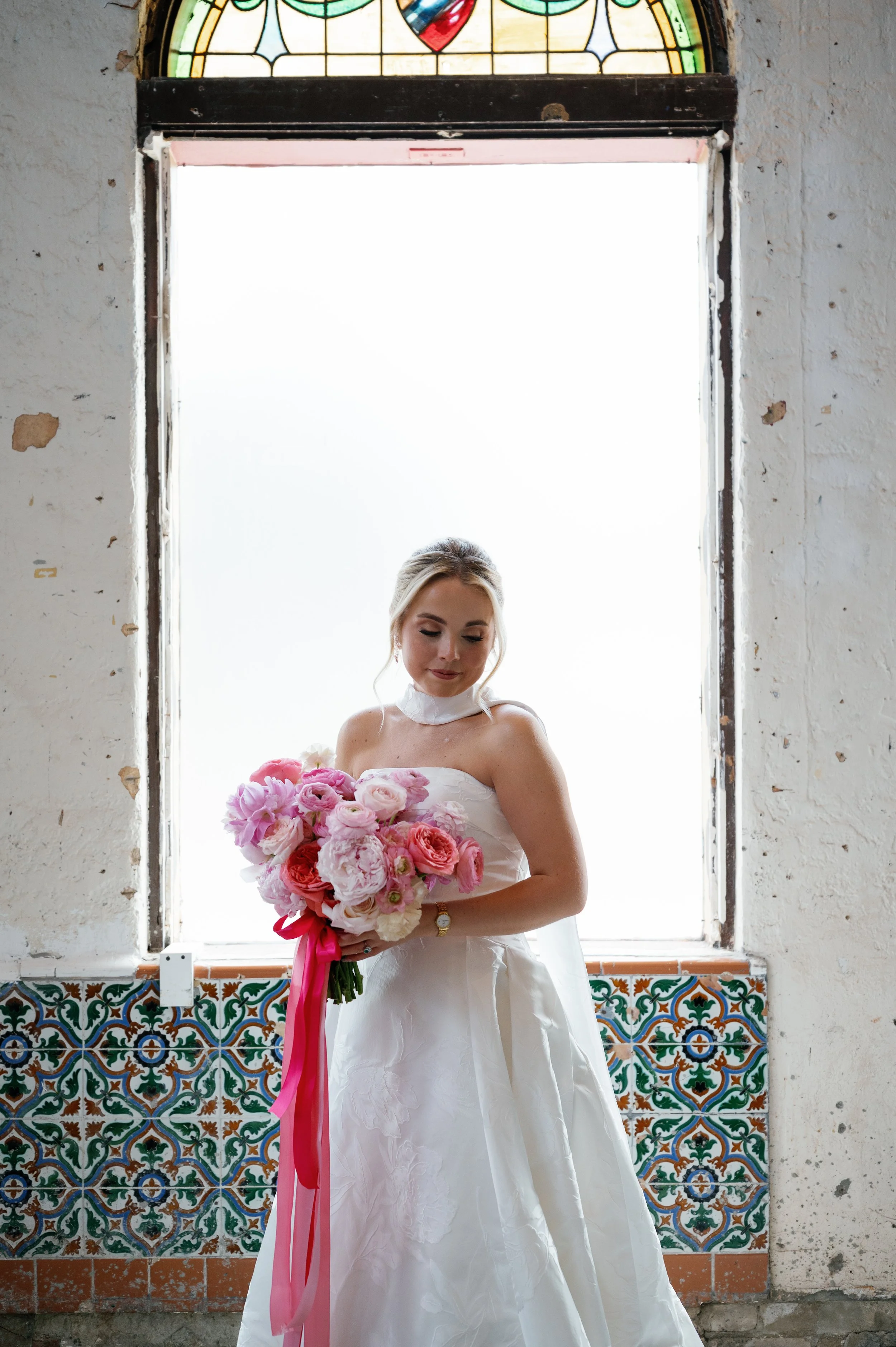 A bride in a white wedding dress holding a large bouquet of pink and white flowers, standing in front of a brightly lit window with stained glass at the top and decorative tiles at the bottom.