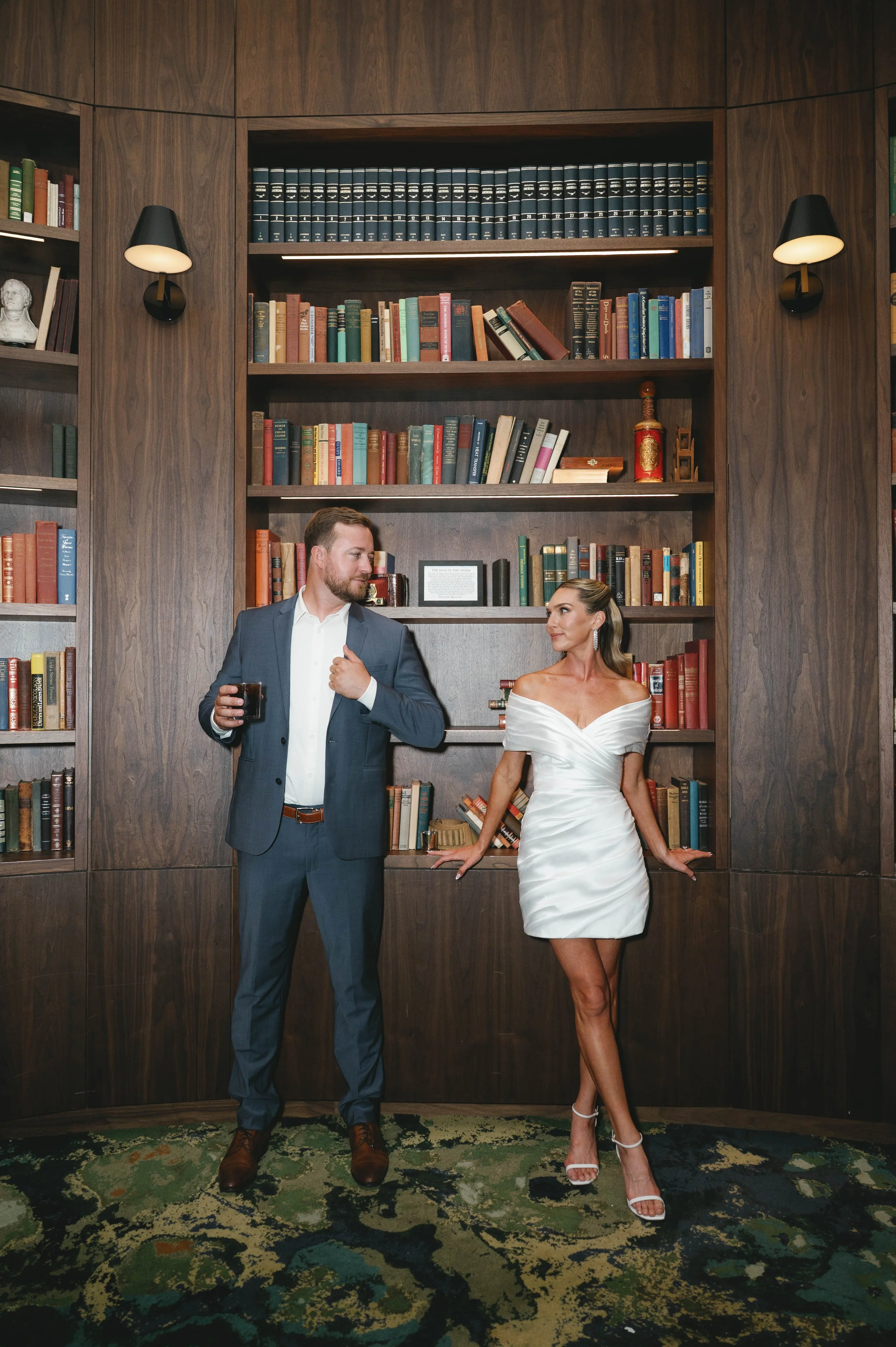 A man in a dark suit holding a drink and a woman in a white dress standing in front of a large wooden bookshelf with multiple shelves filled with books. The room has a patterned carpet and wall-mounted lamps on either side of the bookshelf.