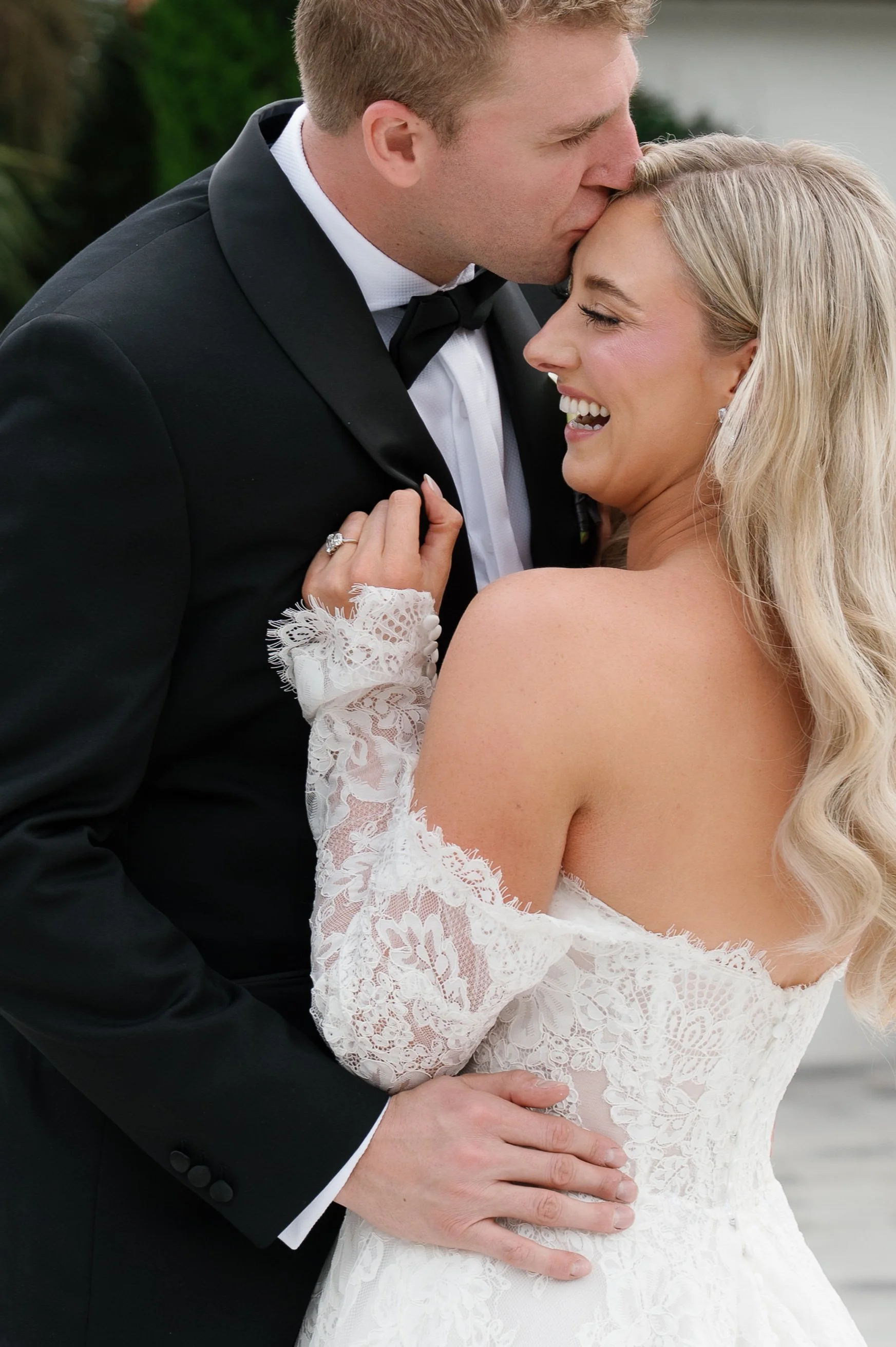A man in a black tuxedo kisses a woman in a white lace wedding dress on the forehead as they embrace outdoors.