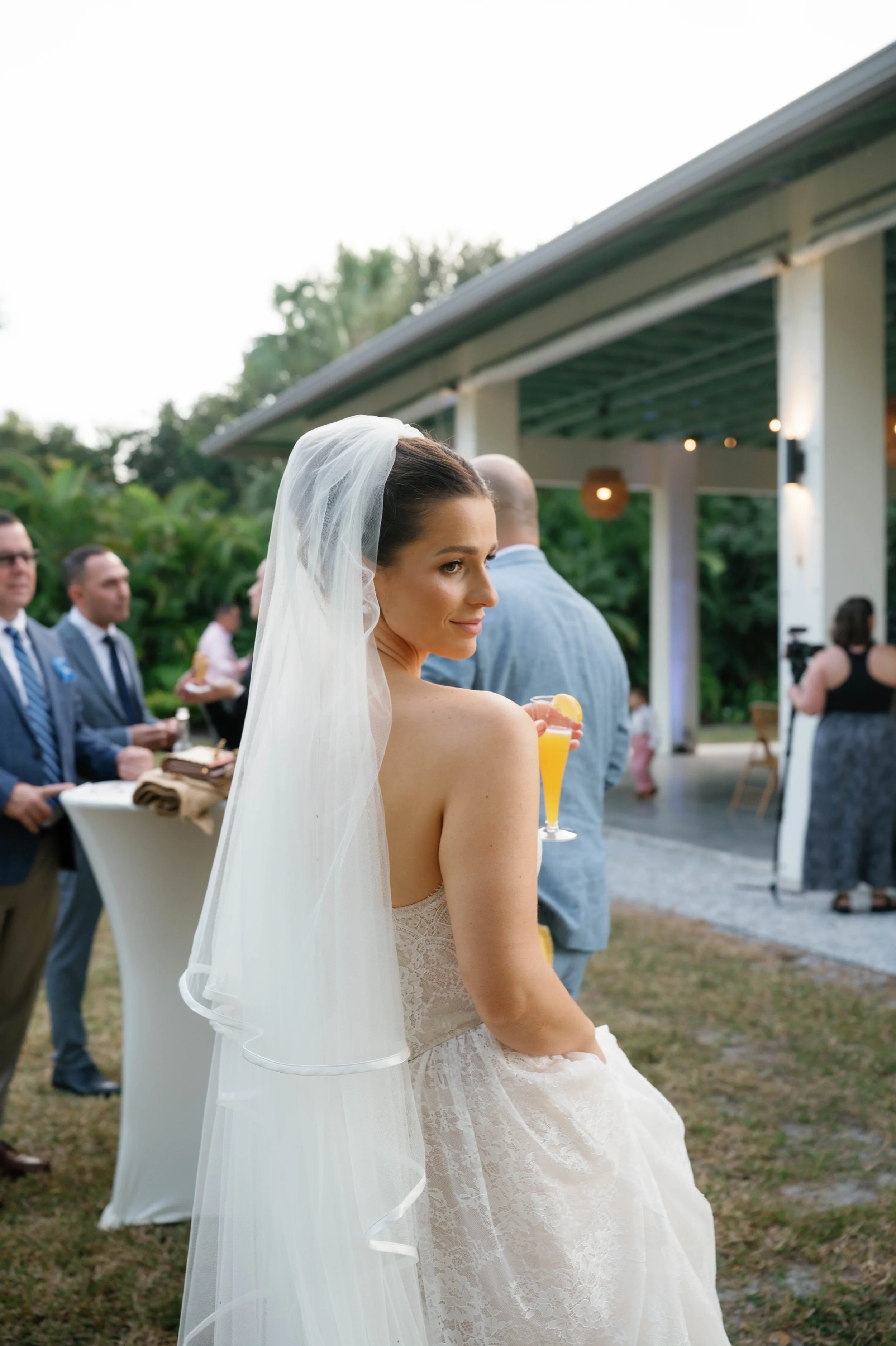 A bride with a veil and strapless lace wedding dress, holding a yellow cocktail with lemon wedge, at an outdoor wedding reception with guests in formal attire.