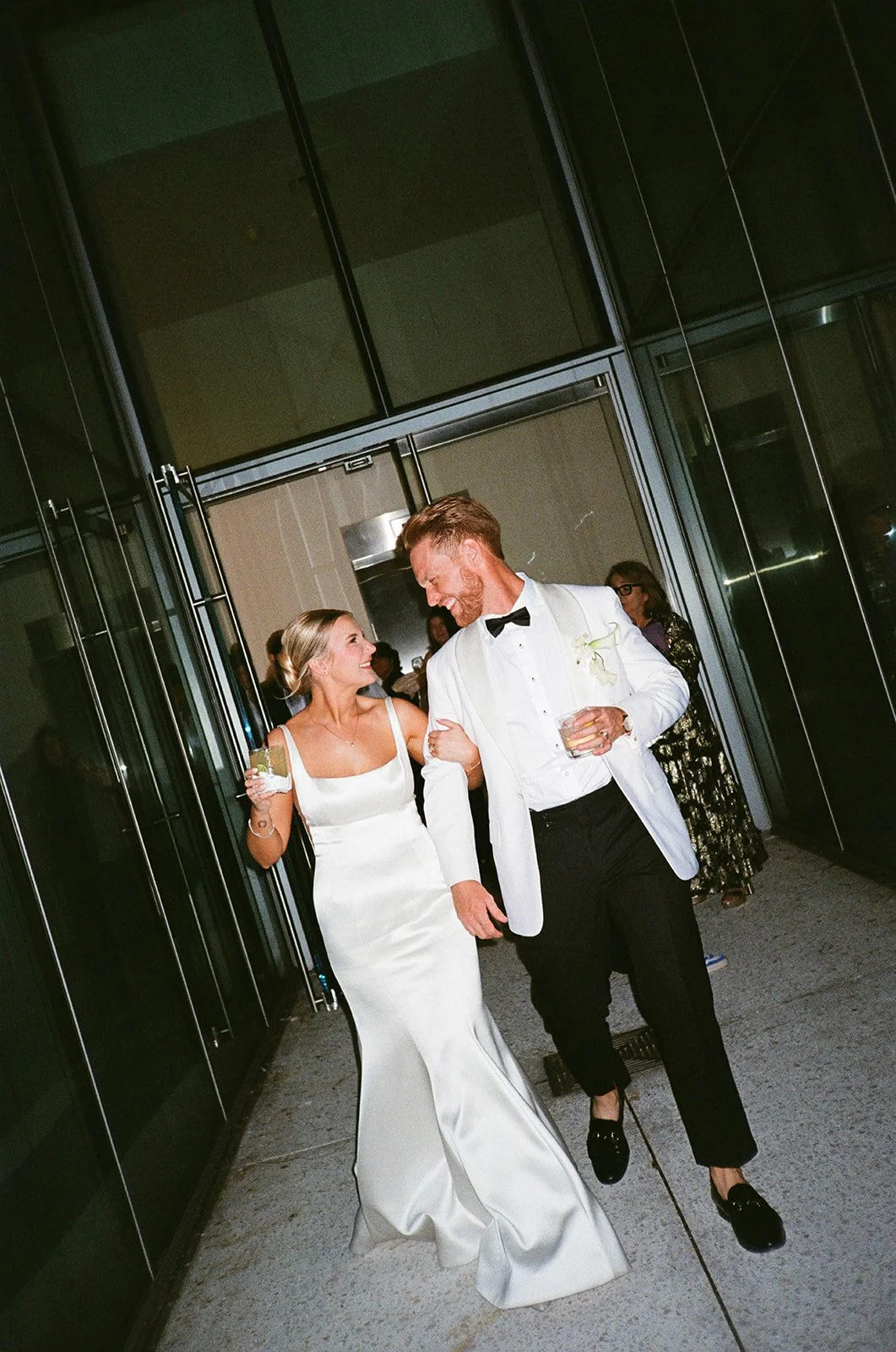A bride and groom walking outside a building, smiling at each other, dressed in wedding attire, holding drinks.