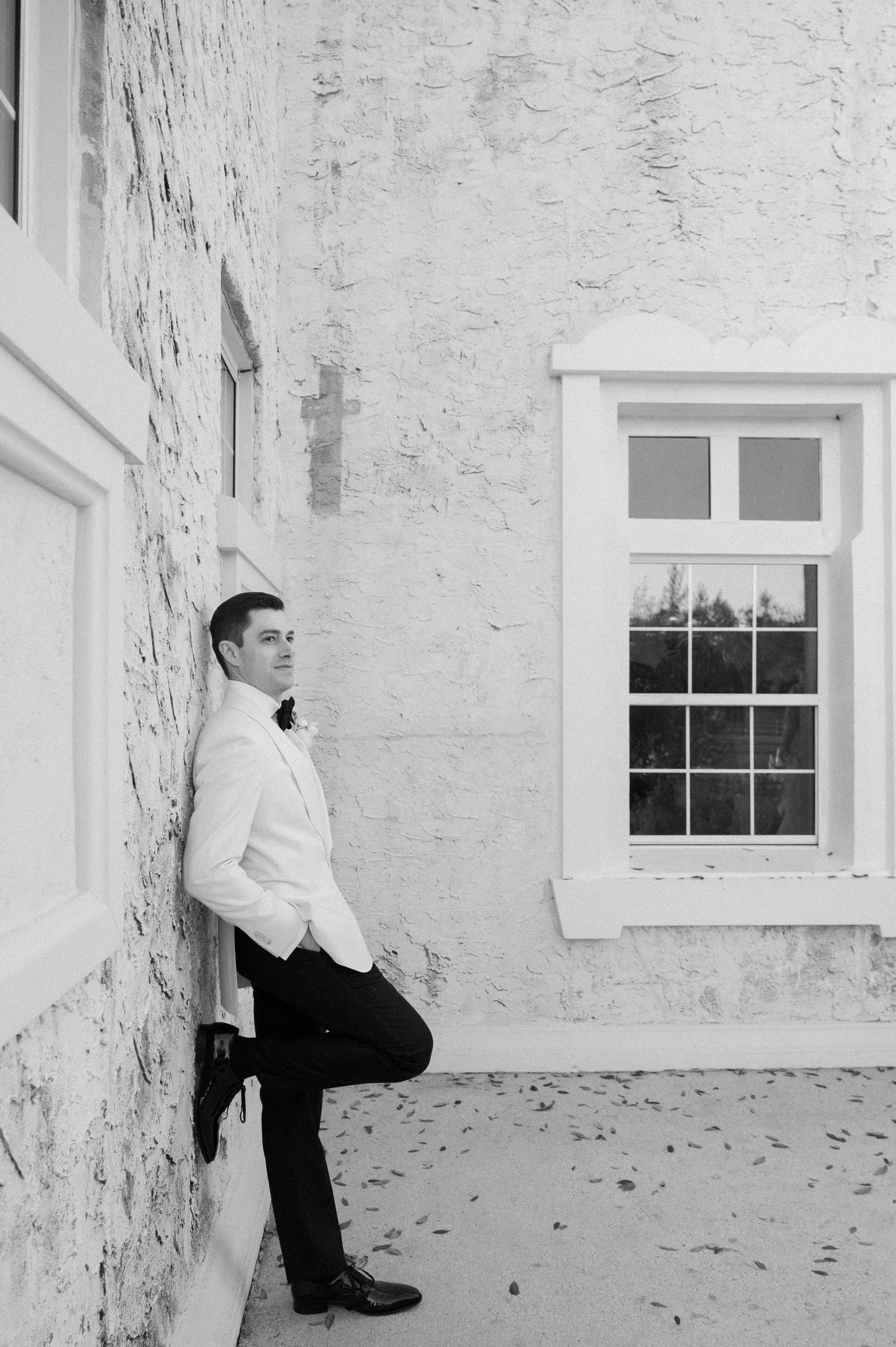 A man in formal attire leaning against a textured brick wall outside a building, with a window to his right and fallen leaves on the ground.