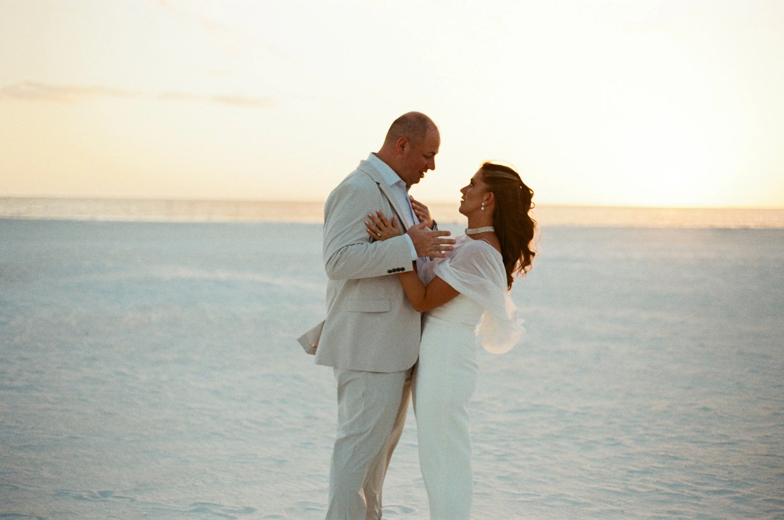A man and woman in wedding attire embrace on a beach at sunset, facing each other closely.