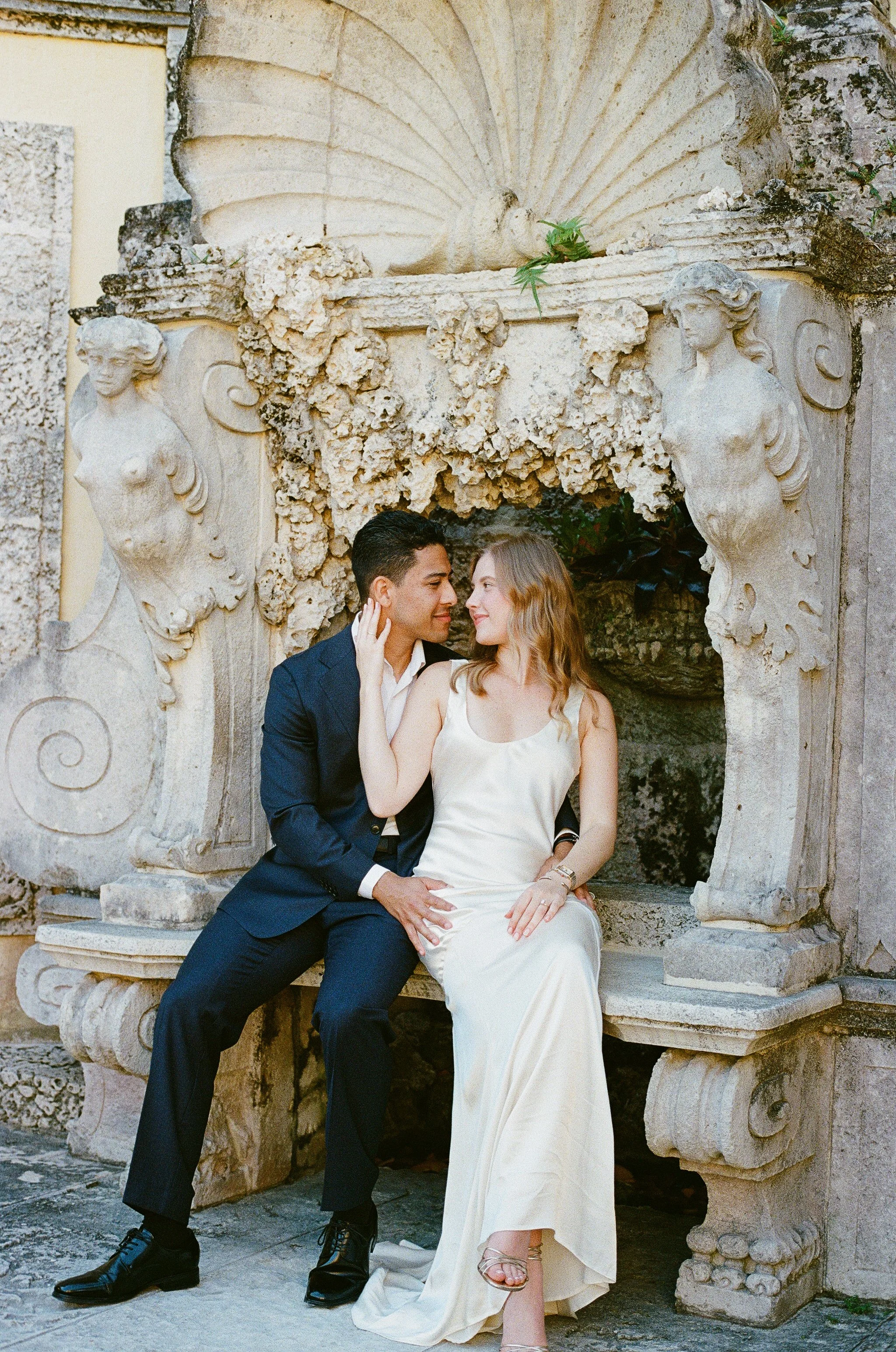 A couple sitting closely together on a carved stone bench with ornate sculptures and carvings, in an outdoor historic or artistic setting.