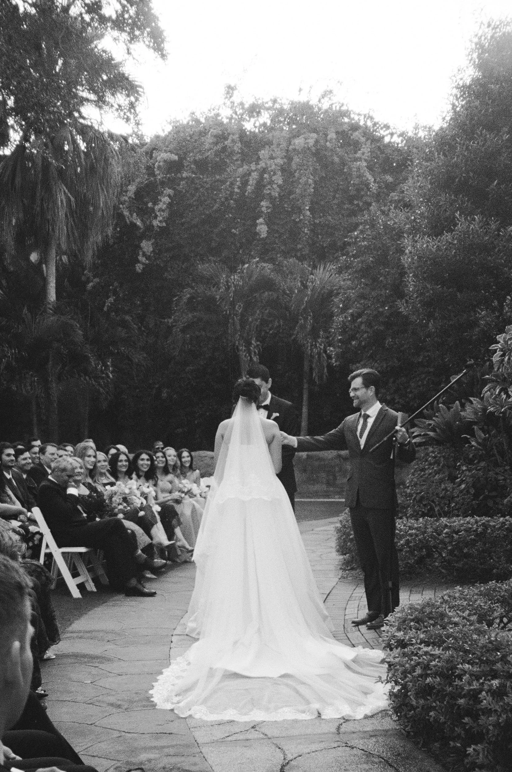 Black and white photo of a wedding ceremony outdoors with guests seated on either side. A bride in a long gown with a train faces two groomsmen, one of whom is holding a microphone and smiling, while the bride and groom stand before him. Lush trees a