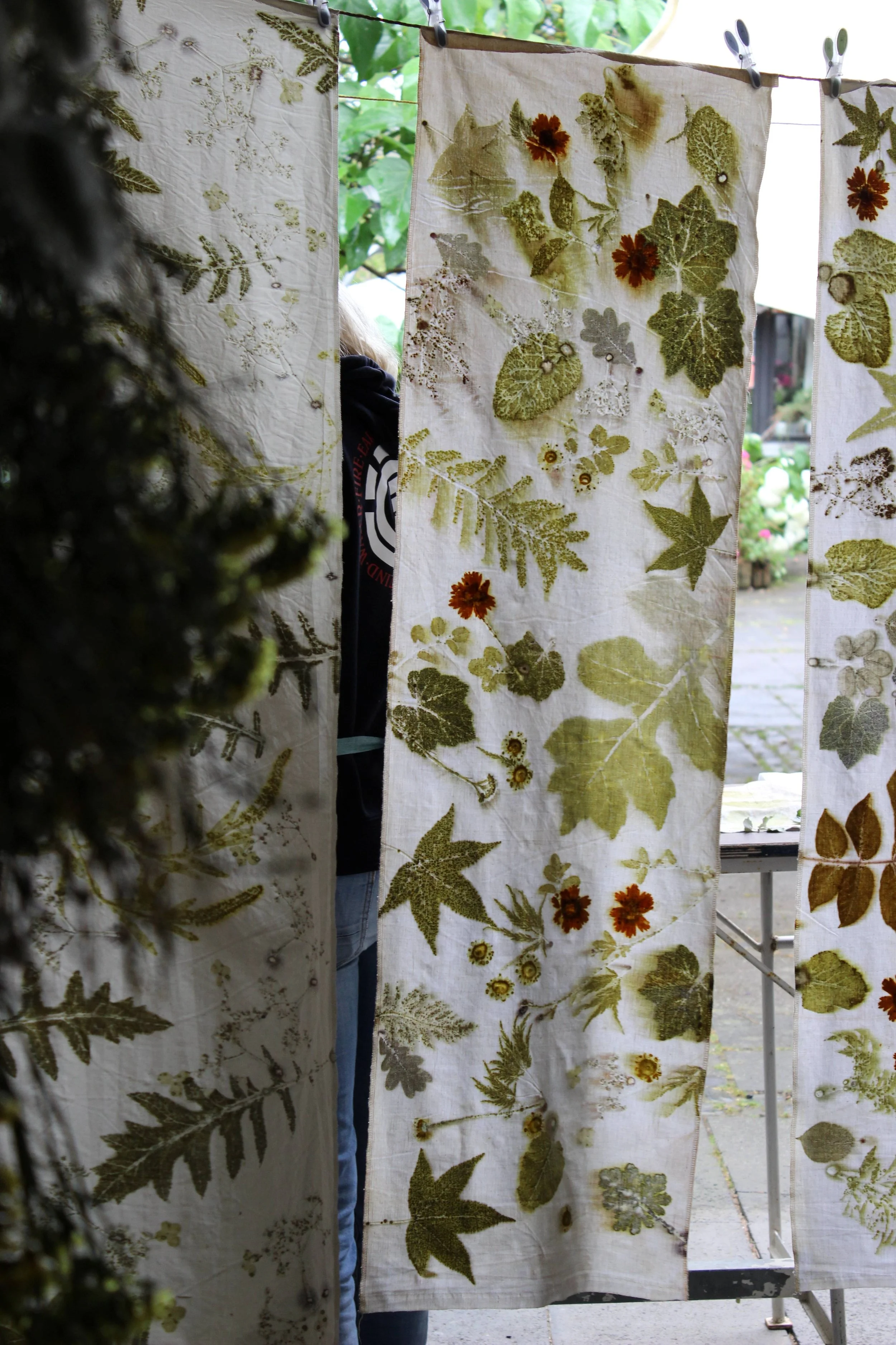 Stoffbahnen mit im Ecoprinting Verfahren aufgedruckten grünen und braunen Blättern und Blumen hängen an einer Leine im Freien.