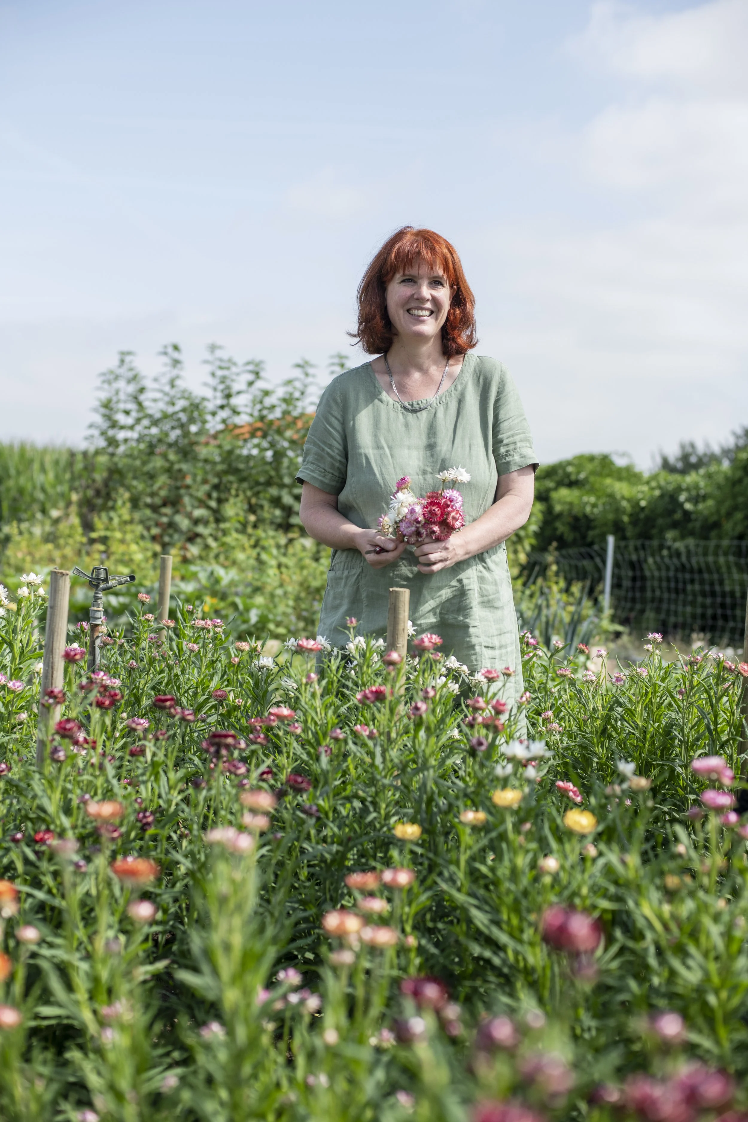 Eine lachende Frau mit roten Haaren in einem grünen Kleid steht in einem Garten mit bunten Blumen, im Hintergrund blauer Himmel mit Wolken.
